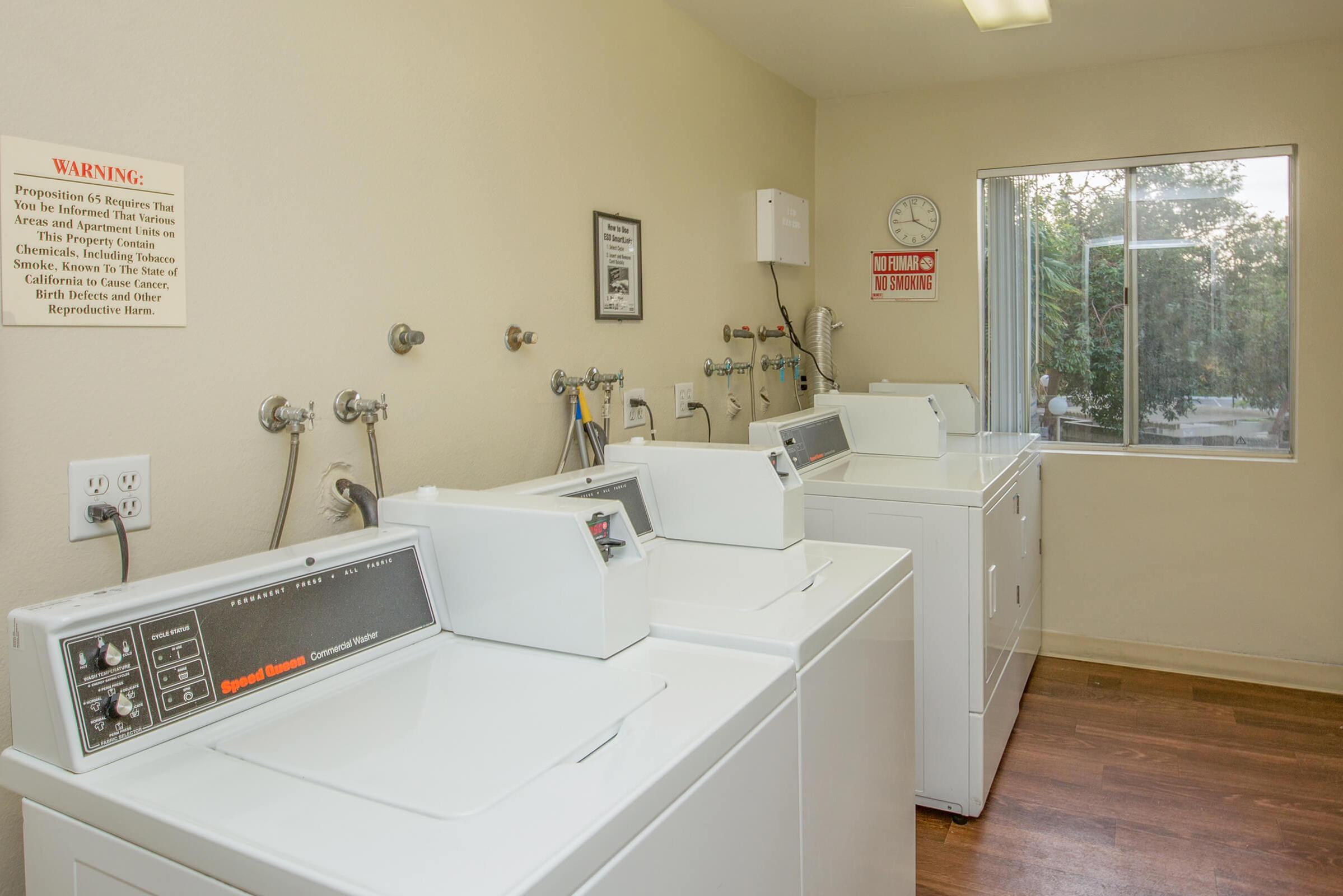 A clean, well-lit laundry room featuring several white washing machines lined up, a window allowing natural light, and wall-mounted utility connections. A clock and a warning sign are visible, adding context to the space. The floor is wooden, giving the room a warm feel.
