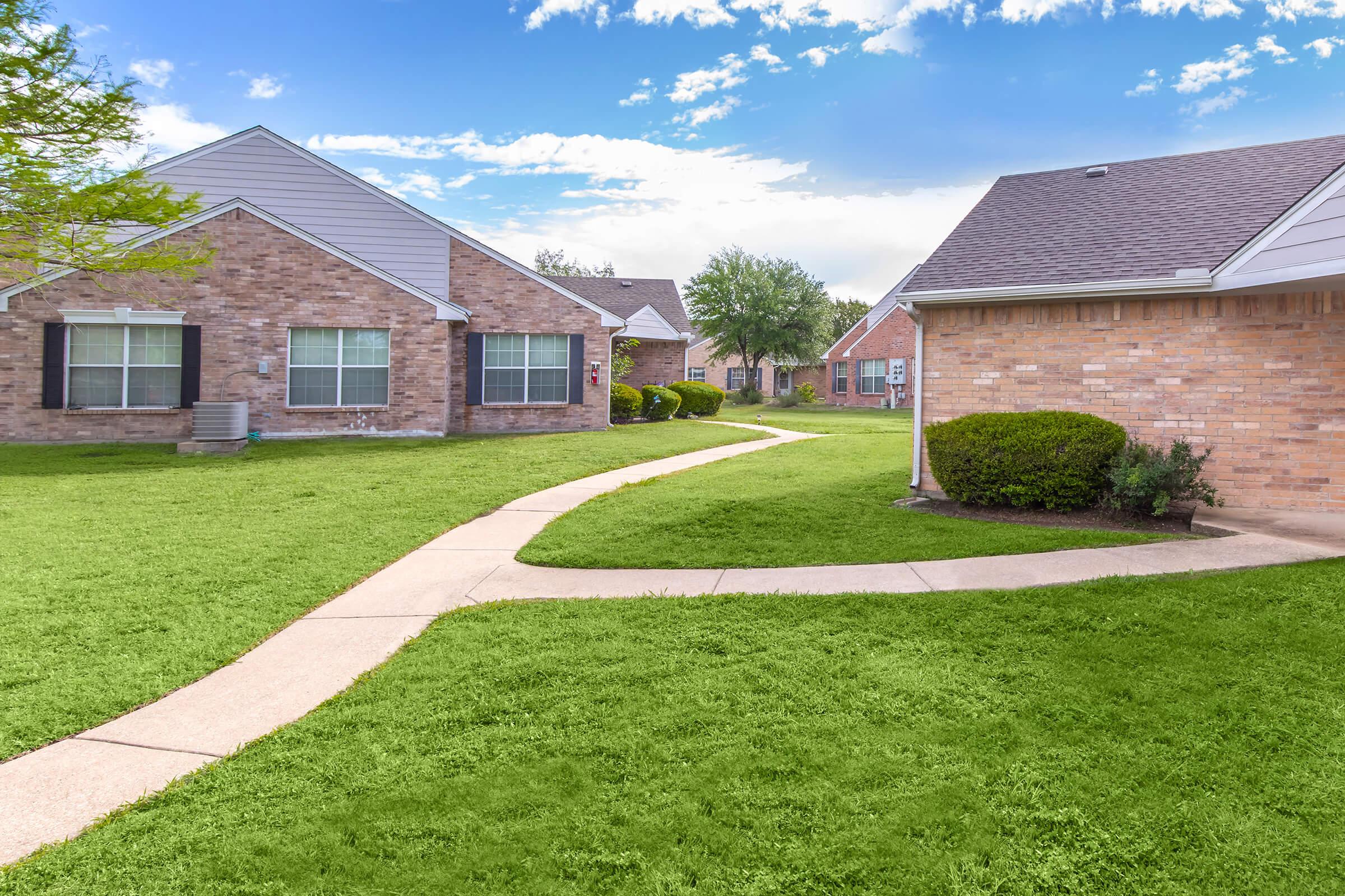 A scenic view of well-maintained residential buildings set in a grassy area. The image features a winding pathway leading through the landscape, flanked by manicured shrubs and trees, under a bright blue sky with scattered clouds. The peaceful environment suggests a welcoming neighborhood atmosphere.