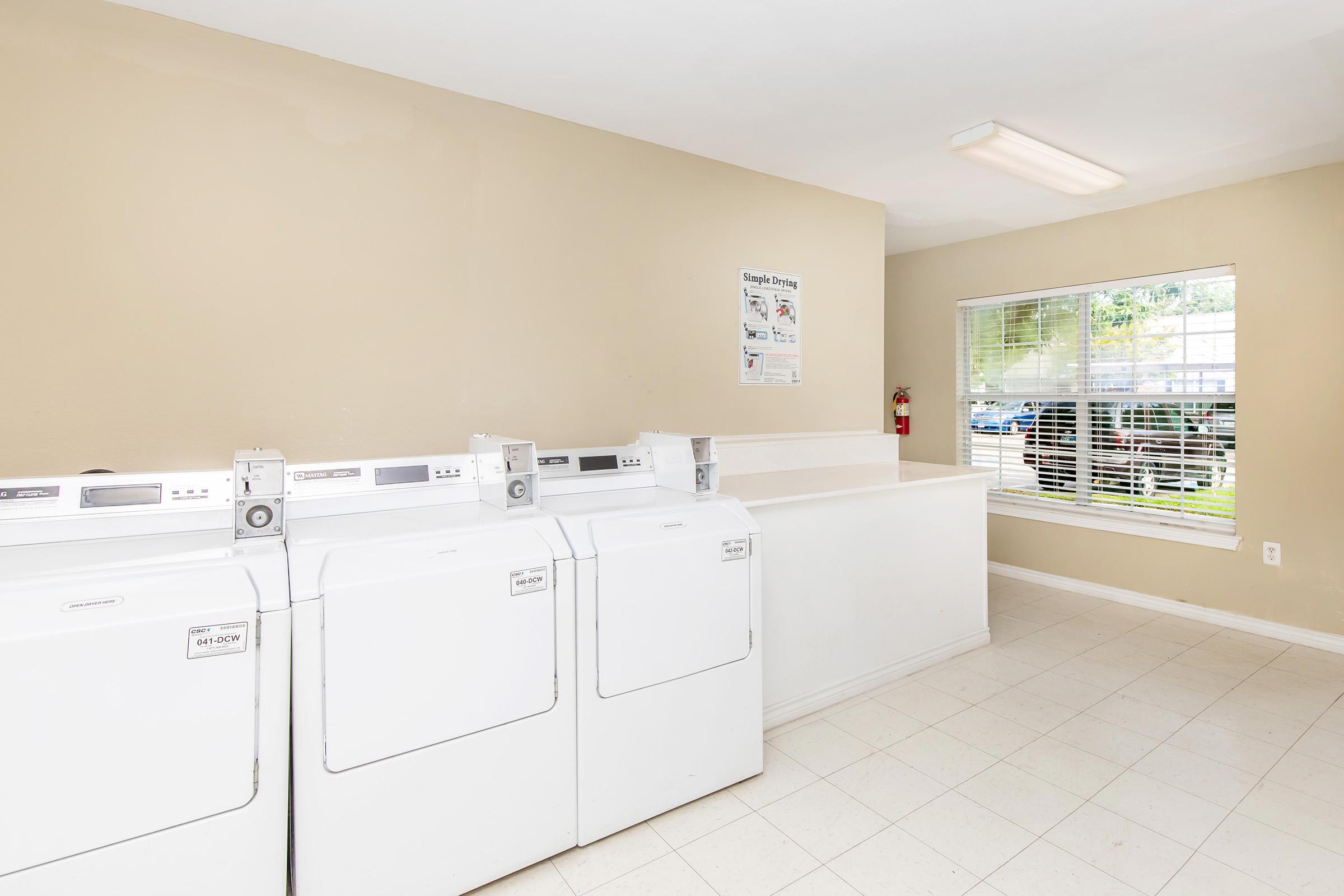 A clean, well-lit laundry room featuring three white dryers along a wall, with a fold-down counter beside them. A window allows natural light into the space, and a fire extinguisher is mounted on the wall. Informational signage is visible above the dryers. The walls are painted in a light beige color.