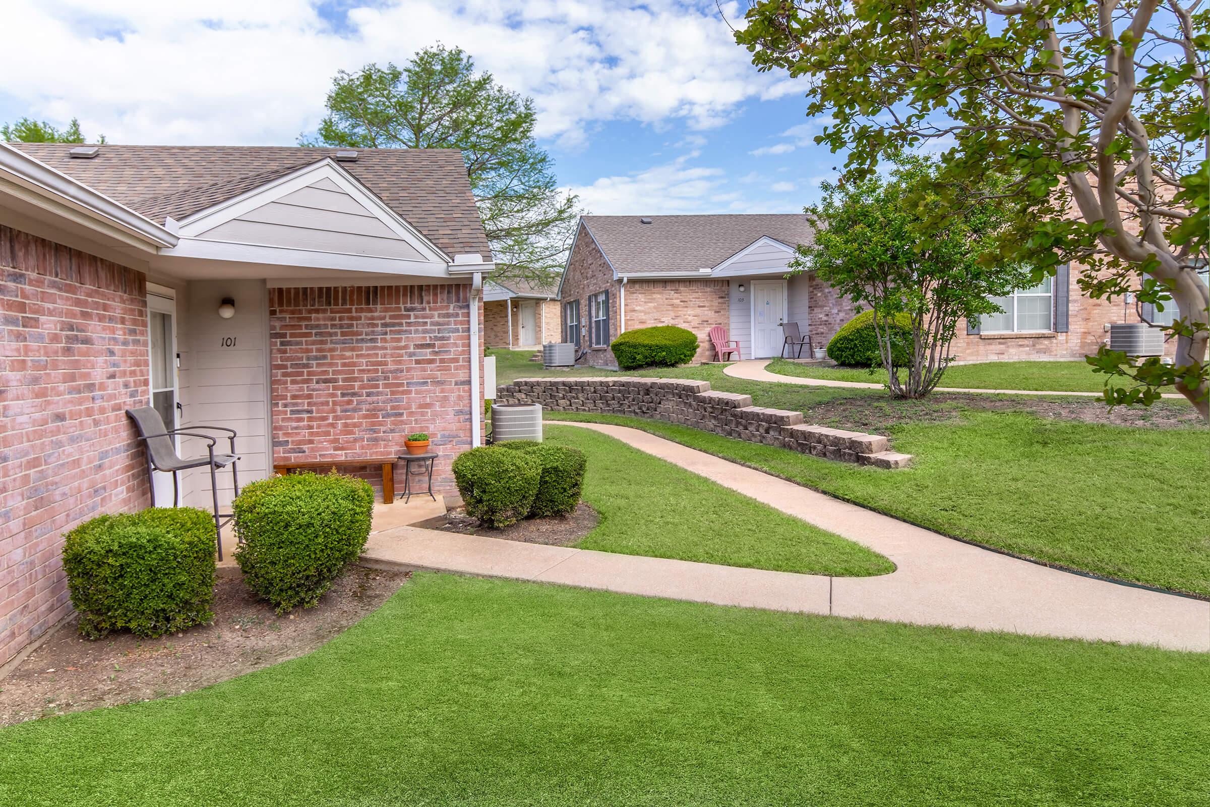 A landscaped view of a residential area featuring brick buildings, a neat pathway, and green grass. A front porch with a chair is visible on the left, while other buildings are in the background. The sky is partly cloudy, adding a serene ambiance to the setting.