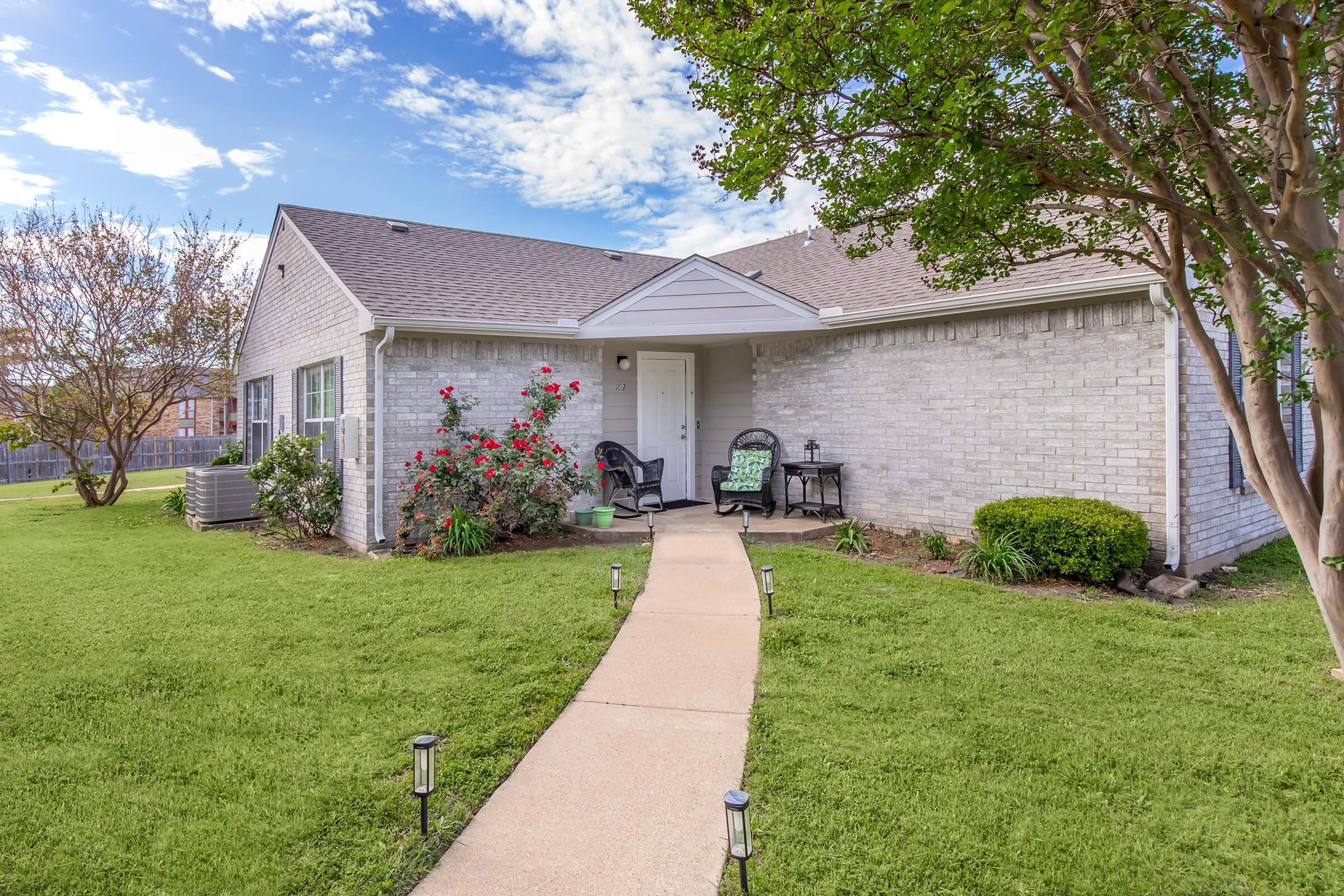 A charming single-story home with a light gray brick exterior surrounded by green grass and blooming rose bushes. A curved walkway leads to the front door, which is flanked by potted plants. Two black chairs with decorative cushions sit on a small porch, under a clear blue sky with scattered clouds.