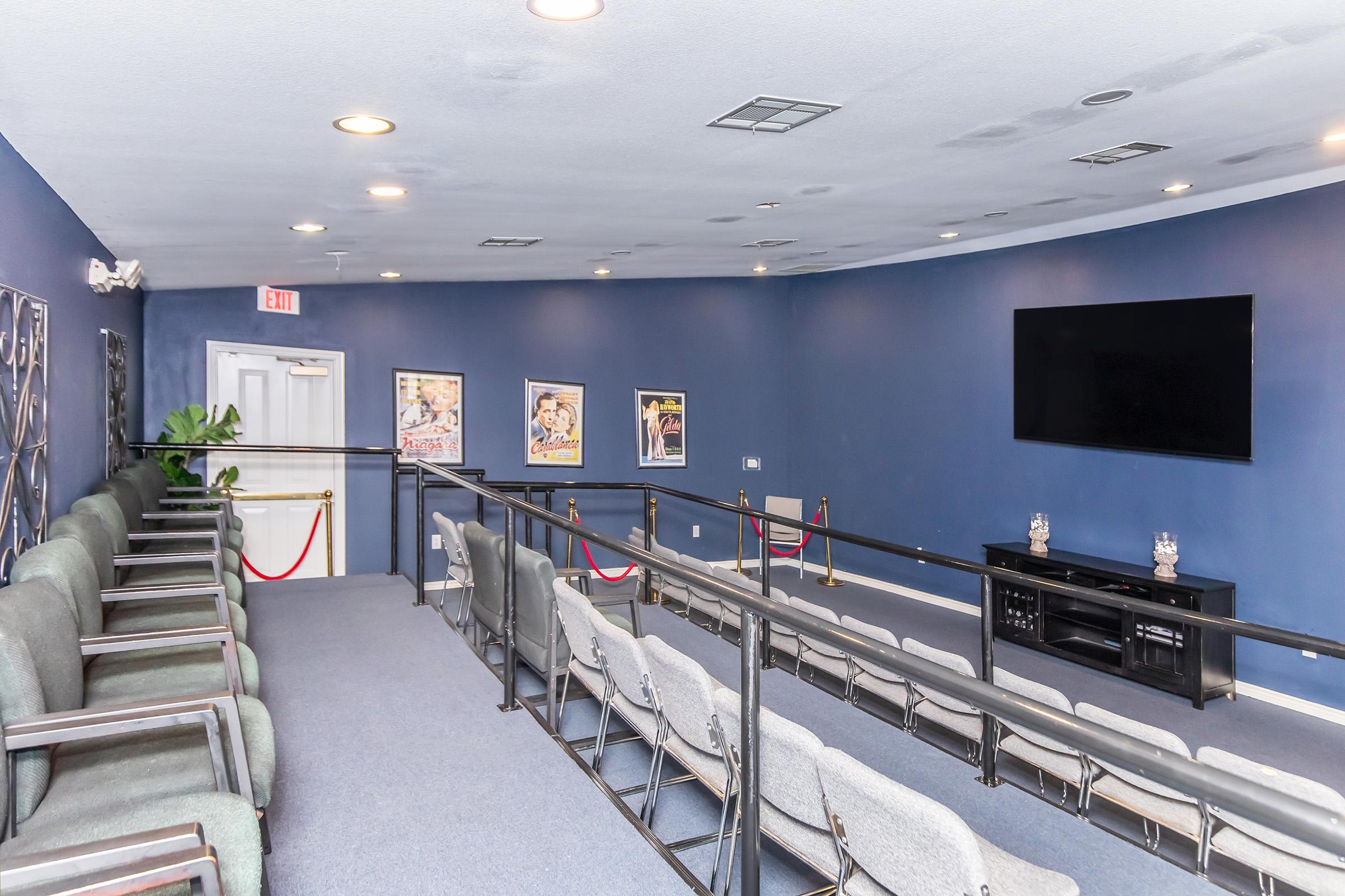 Interior view of an audience seating area with gray chairs arranged in rows facing a wall-mounted TV. The walls are painted dark blue and feature framed movie posters. A railing separates the seating from an empty section, and there are red ropes indicating a reserved area. The overall atmosphere is calm and organized.
