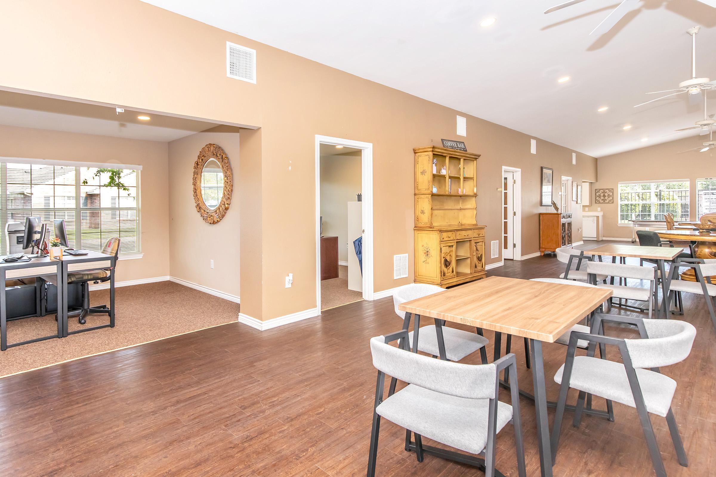 Spacious interior view of a modern living area featuring wooden flooring, a dining table with grey chairs, and a cozy seating area. A desk setup is visible in a separate section, along with a large decorative mirror and a wooden cabinet. The room is well-lit with natural light.