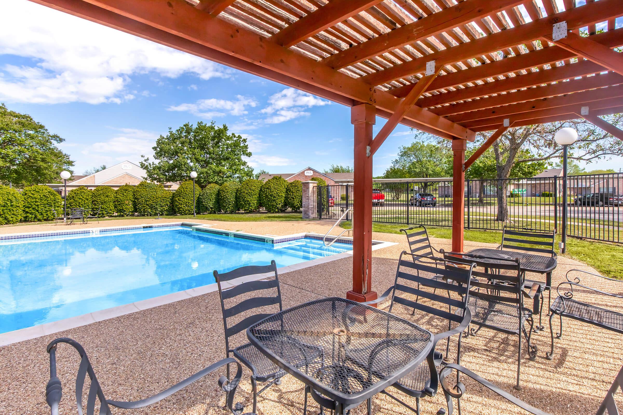 A sunny outdoor space featuring a swimming pool surrounded by a patio with black wrought iron tables and chairs. There is a wooden pergola providing shade, lush green trees in the background, and a fenced area with manicured shrubs.