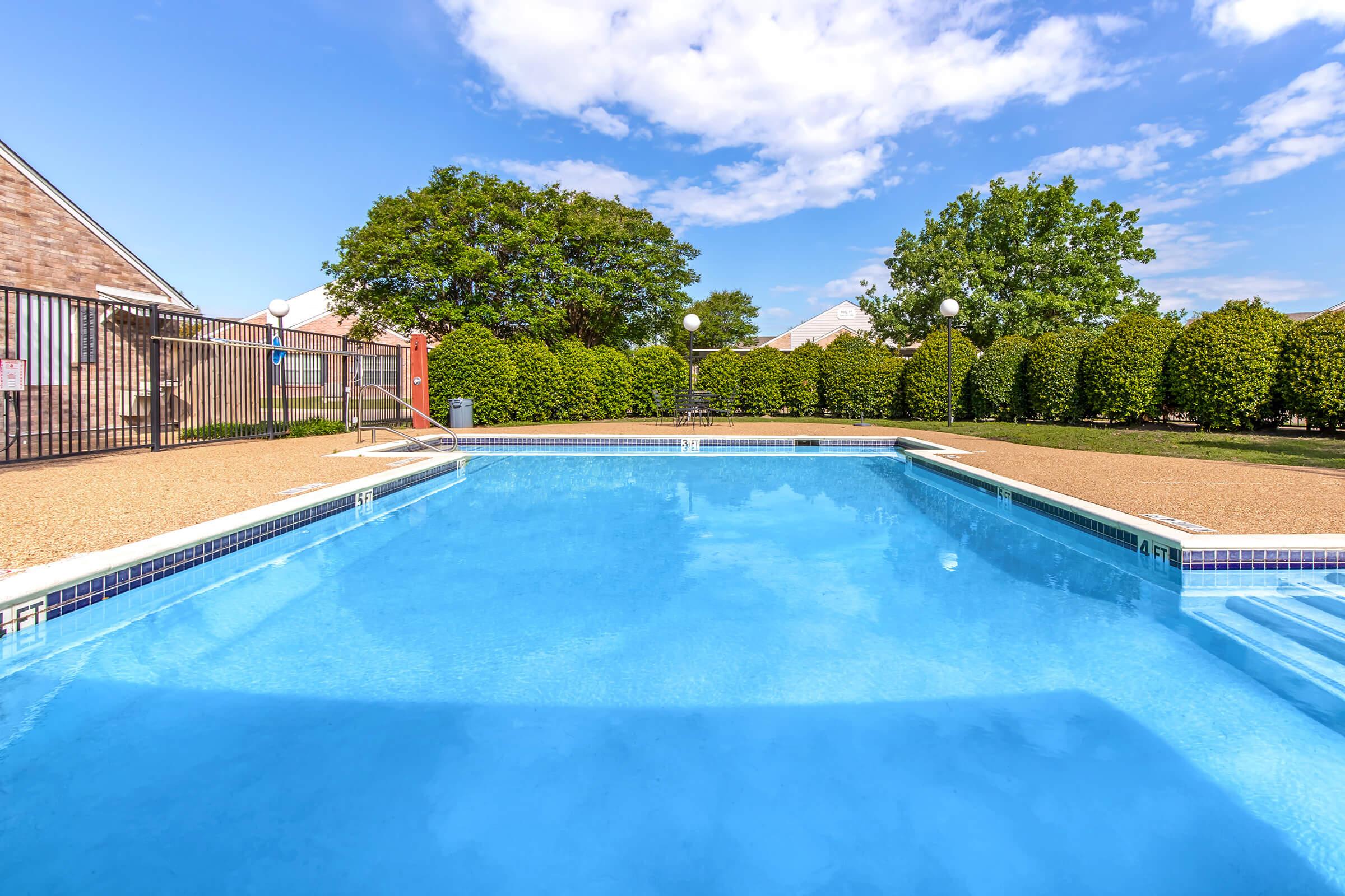A clear blue swimming pool surrounded by a tan deck, with a green hedge and trees in the background. The sky is bright with scattered clouds, and there are light fixtures along the pool area. A fence is visible in the background, providing a sense of privacy.