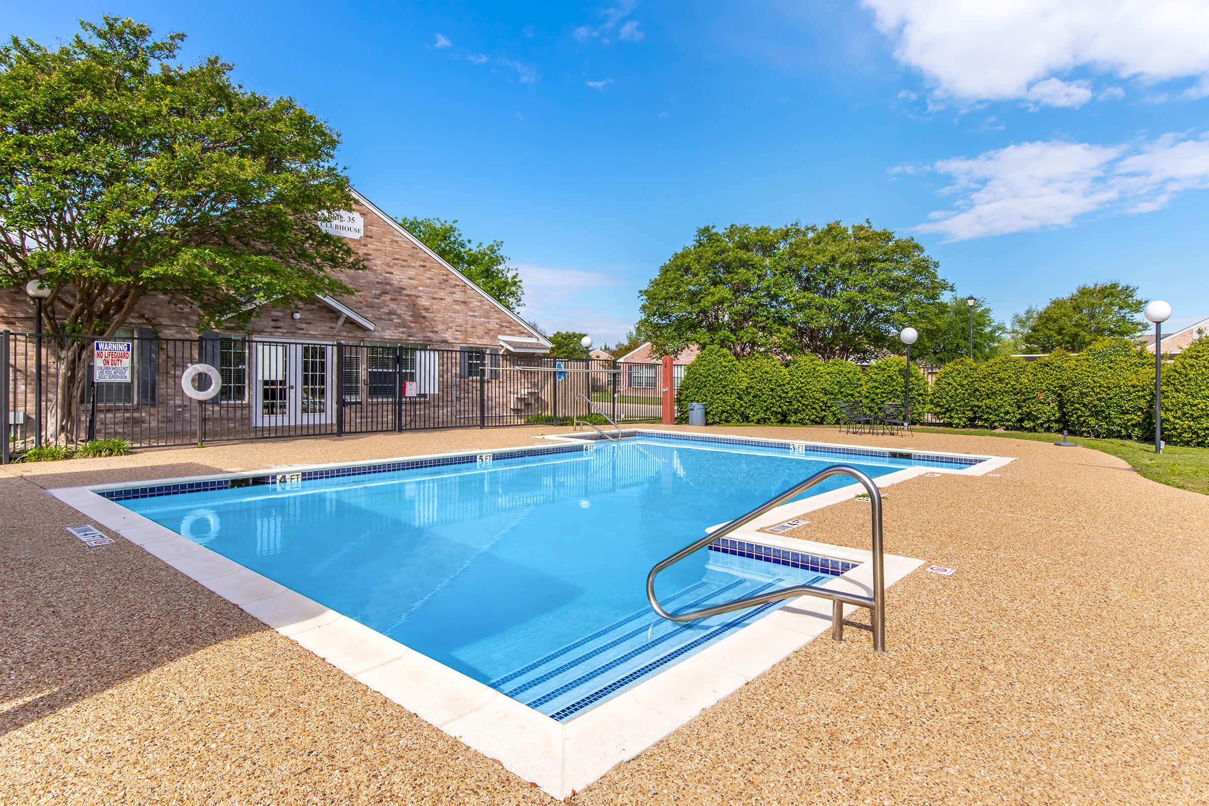 A clean, inviting outdoor swimming pool surrounded by a textured stone deck. Lush green trees and shrubs line the background, while a charming building with a brick facade is visible nearby. Clear blue sky and bright clouds enhance the vibrant atmosphere of the scene.