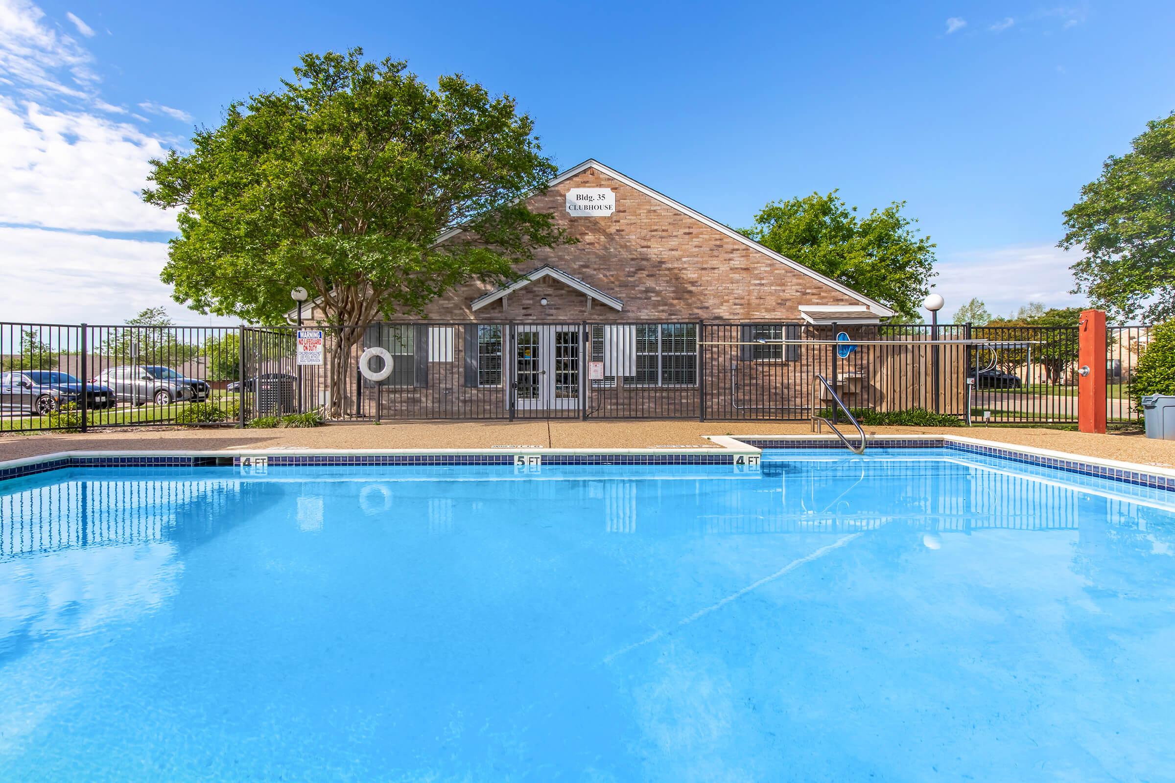 A clear blue swimming pool with steps leading into the water, surrounded by a tiled deck. In the background, there is a brick building with large windows and a sign. Green trees provide shade around the pool area under a bright blue sky with scattered clouds. A safety ring is visible on the poolside.