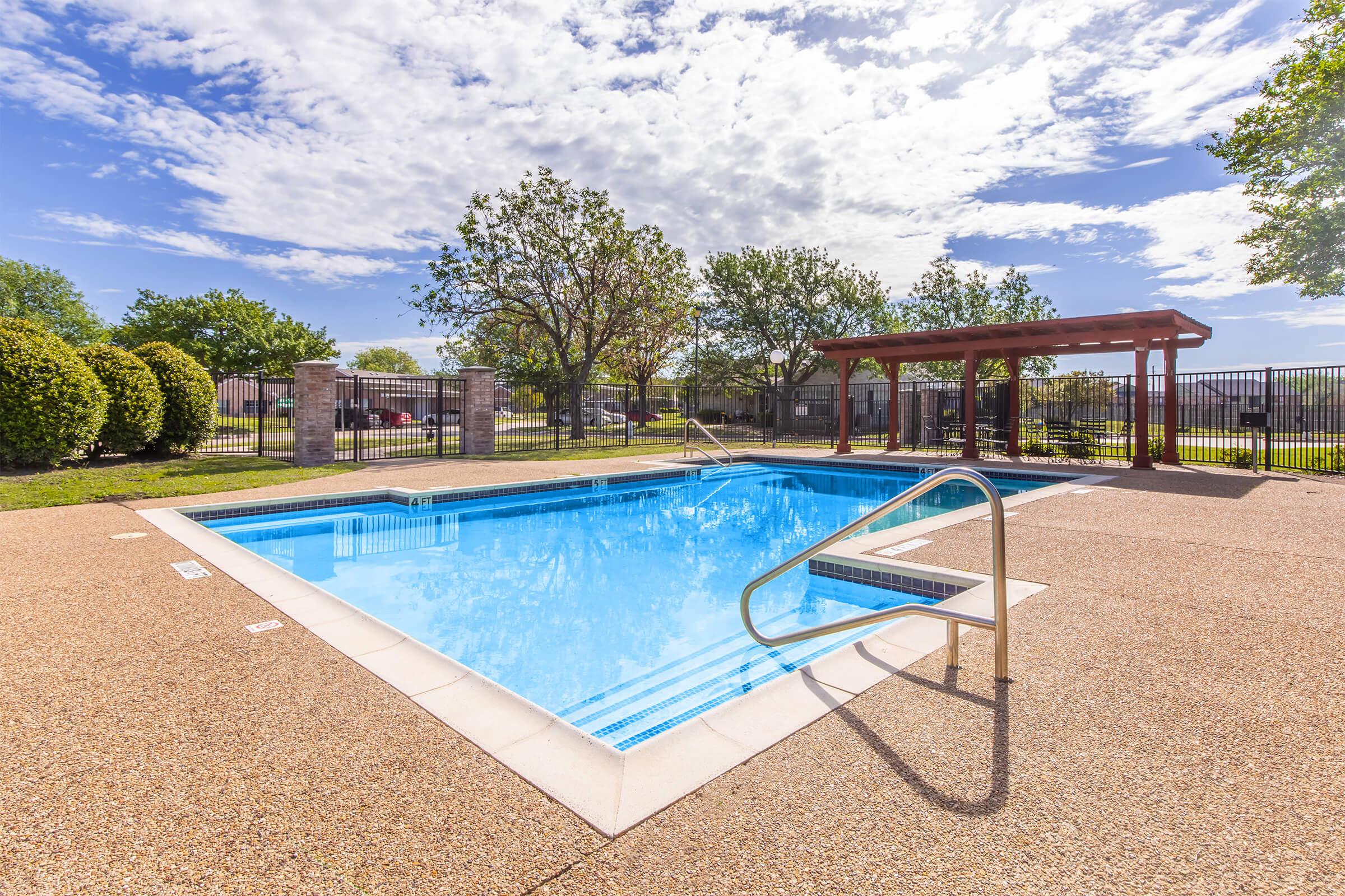 A bright outdoor swimming pool surrounded by a paved deck, featuring a ladder for access. Lush green trees and shrubs frame the area, and a wooden pergola provides shade. The sky is clear with fluffy clouds, creating a vibrant atmosphere for relaxation and leisure.