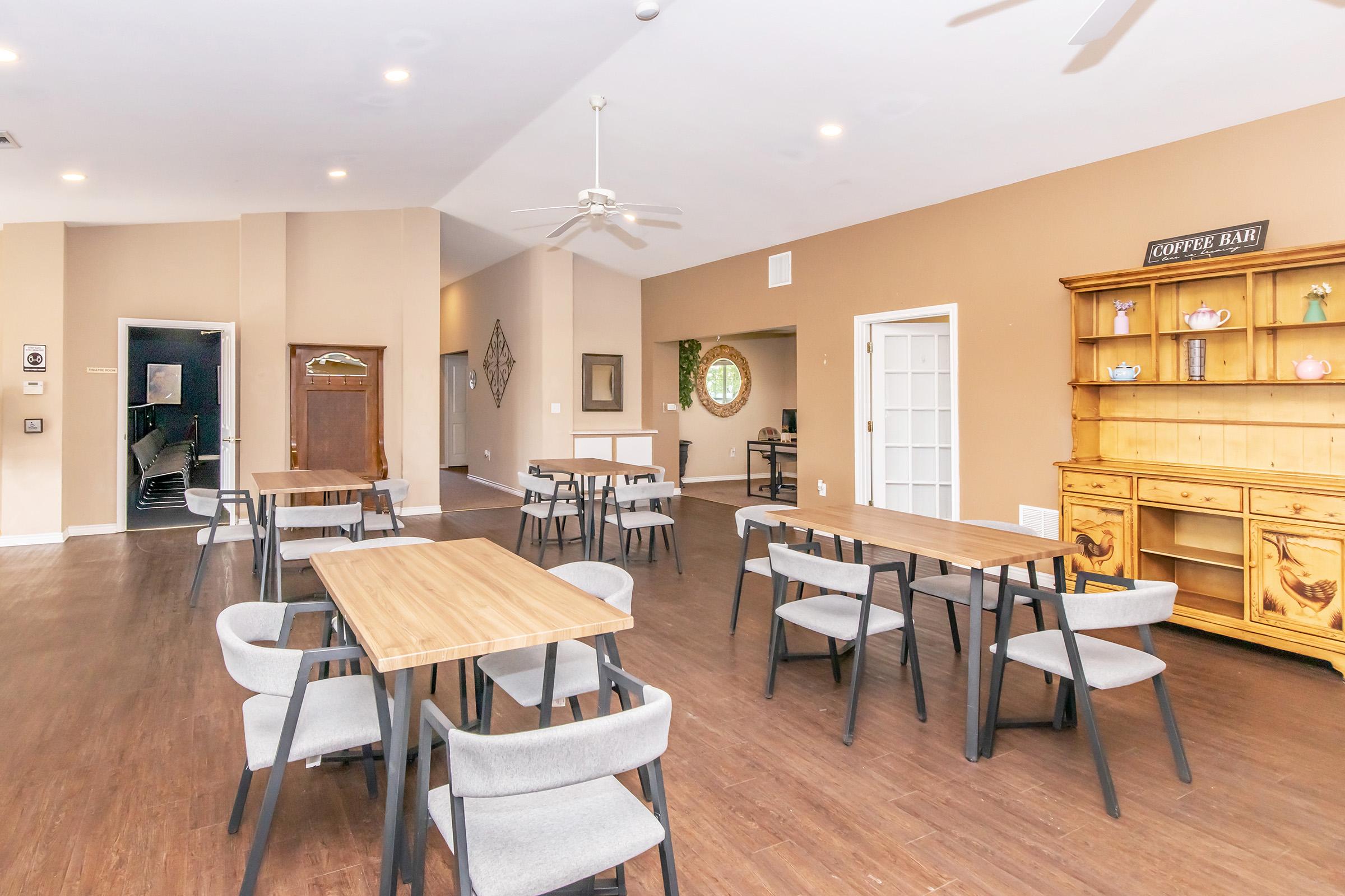 A spacious indoor setting with several wooden tables and chairs, a large wooden cabinet, and a decorative mirror on the wall. The walls are painted in warm tones, and overhead lighting brightens the area. A doorway leads to another room in the background.