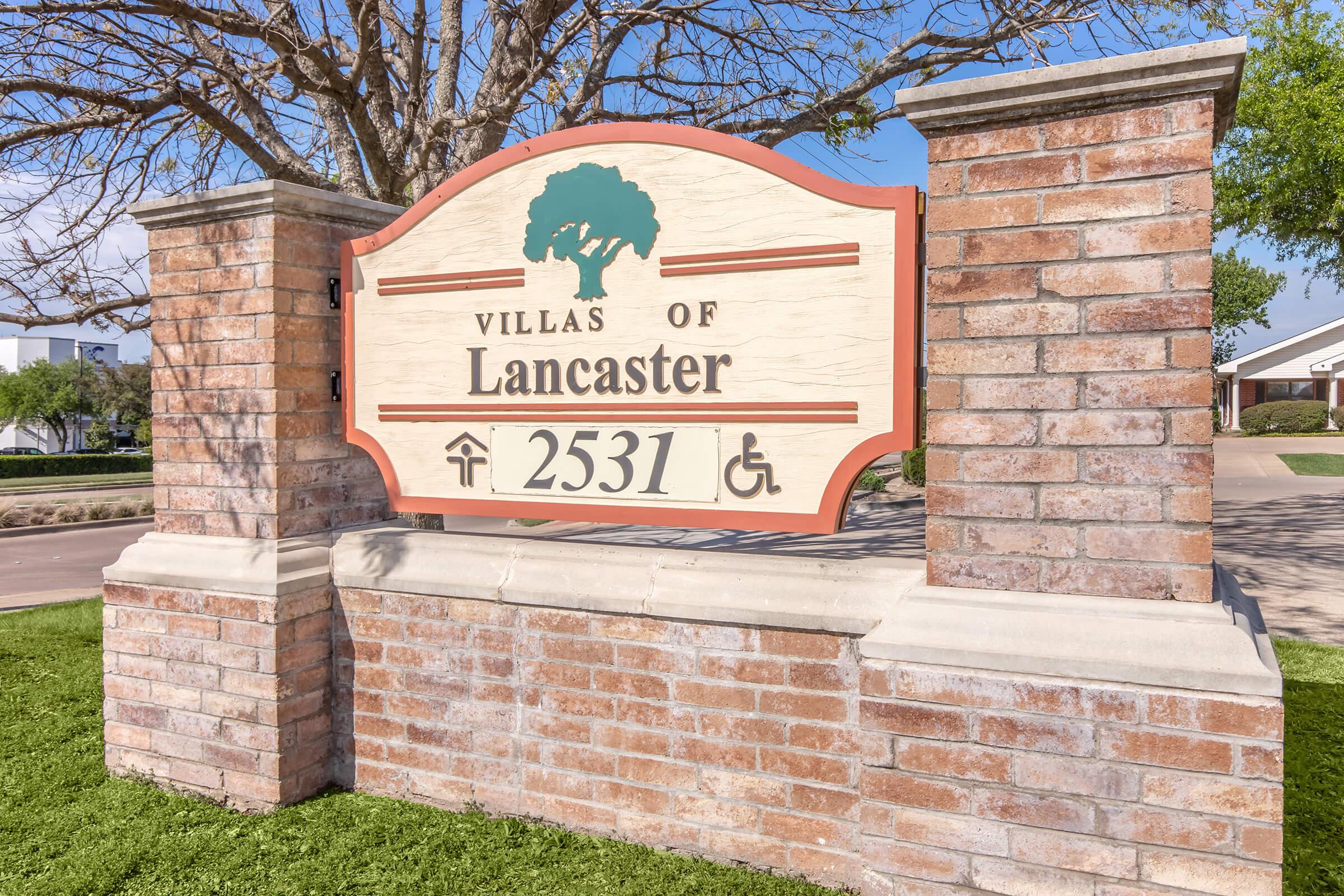 Sign for "Villas of Lancaster" with the address "2531." The sign is mounted on a brick structure surrounded by green grass and a clear blue sky in the background. Accessible symbols are displayed, indicating wheelchair access.