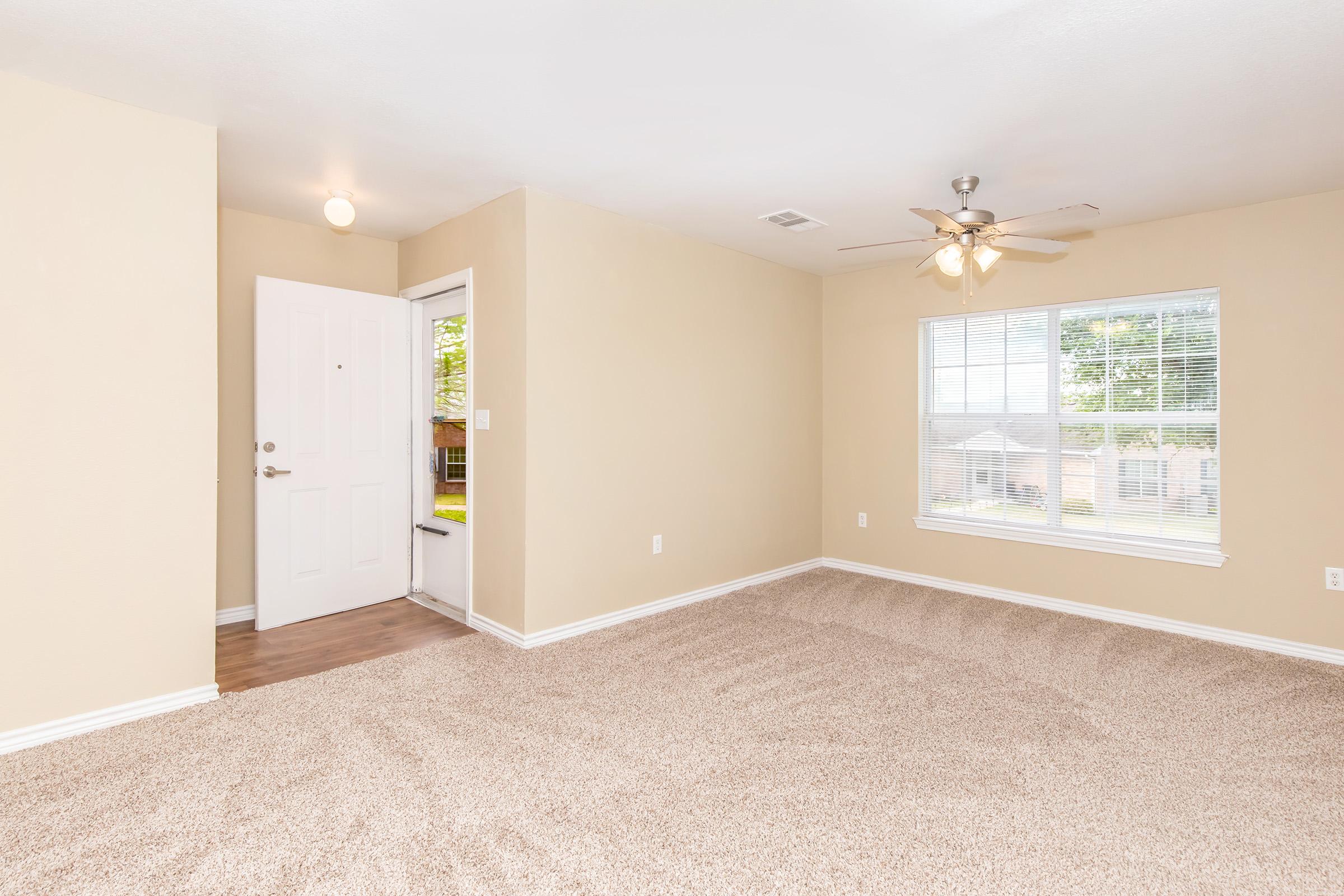 A well-lit room with beige walls and a light brown carpet. It features a white front door, a window with blinds, and a ceiling fan. The space appears empty and inviting, with natural light coming in from the window. The layout is open, creating a cozy atmosphere.