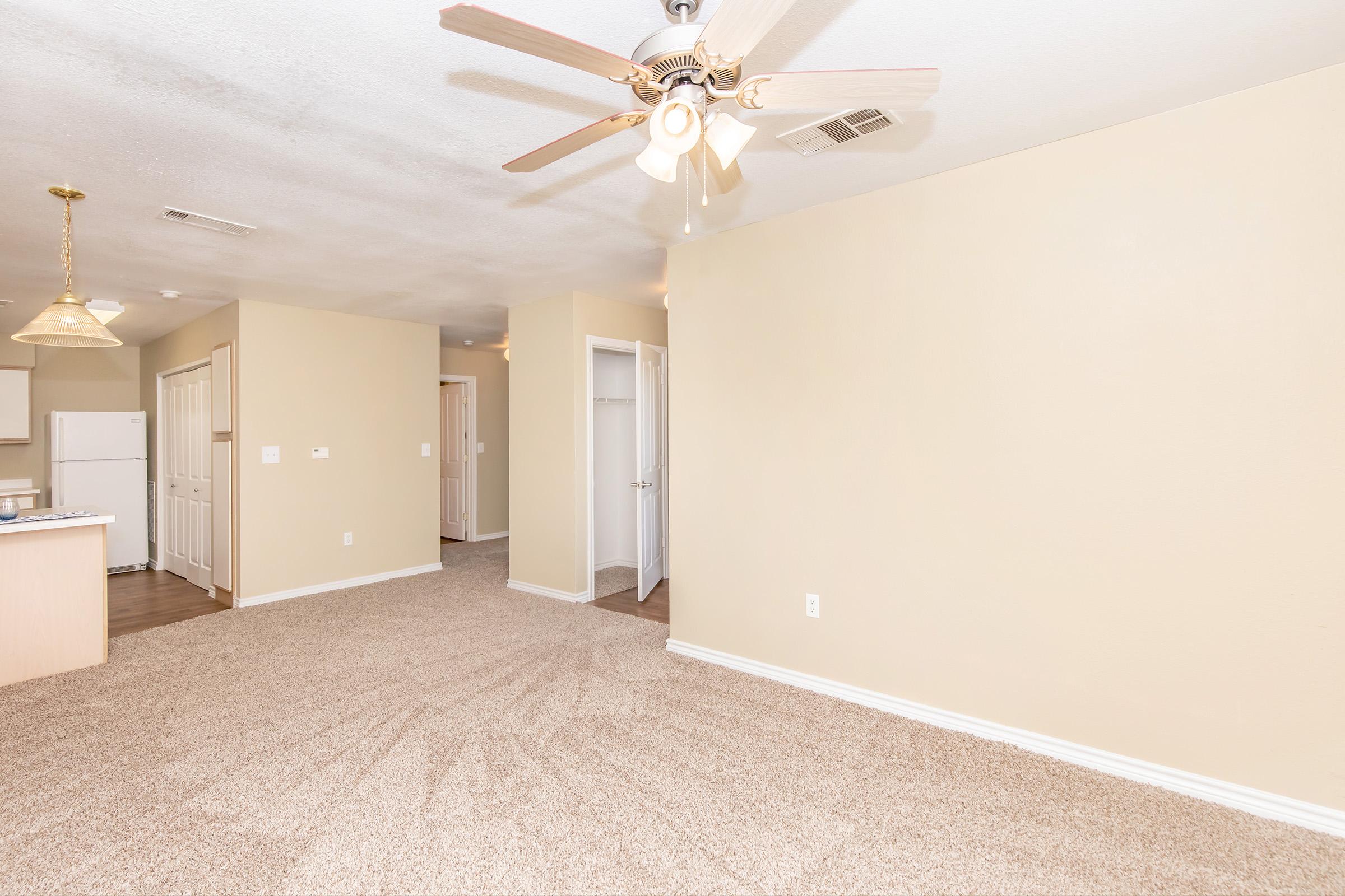 A spacious room with light beige walls and carpet. A ceiling fan is visible, and there is a doorway leading to another room. The kitchen area includes a refrigerator and counter space. Natural light illuminates the space, creating a warm atmosphere.