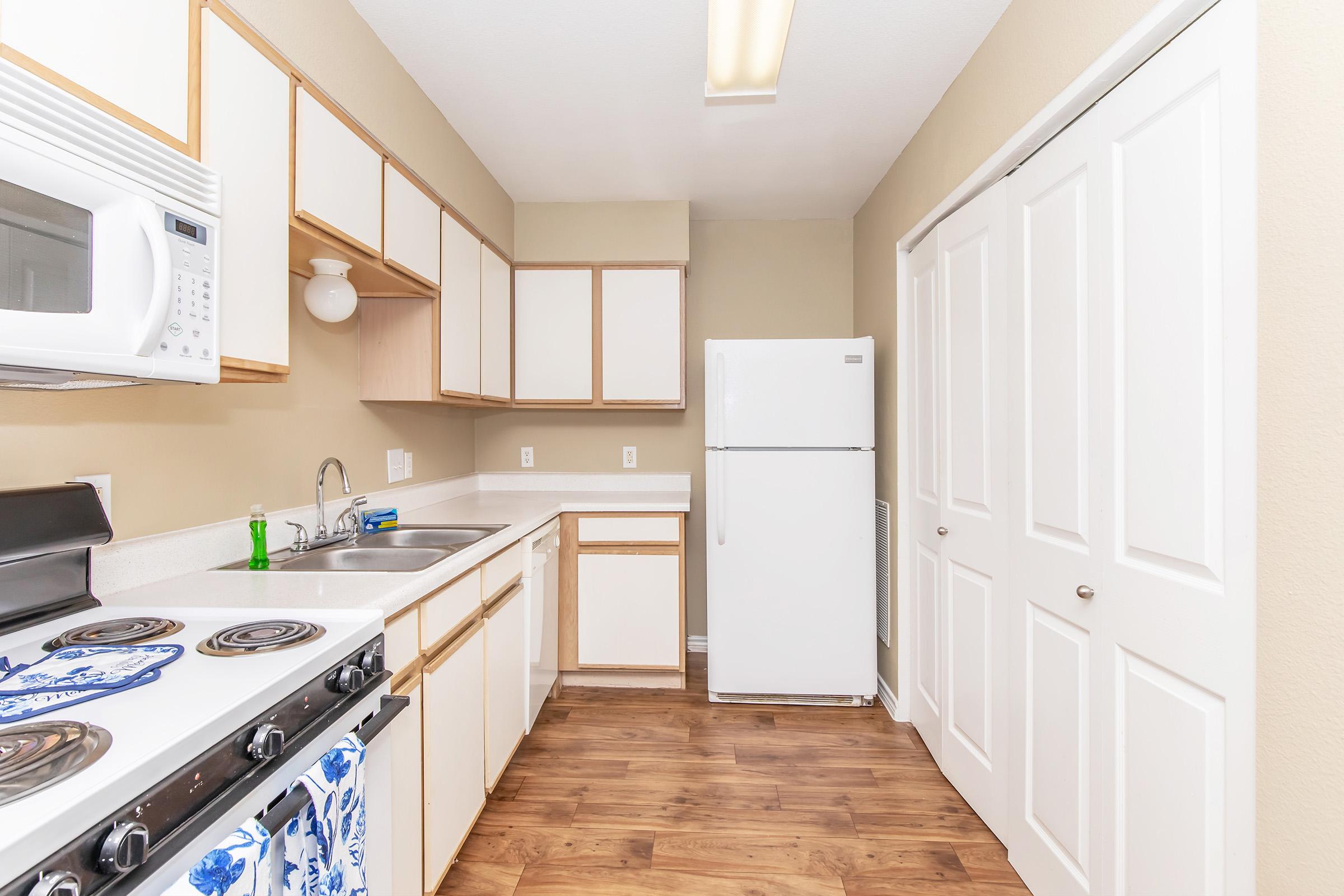 A modern kitchen featuring white cabinets, a microwave, a stove with a blue decorative towel, a double sink, and a white refrigerator. The walls are painted a light beige, and there are closet doors visible on the right side, with wood flooring throughout.