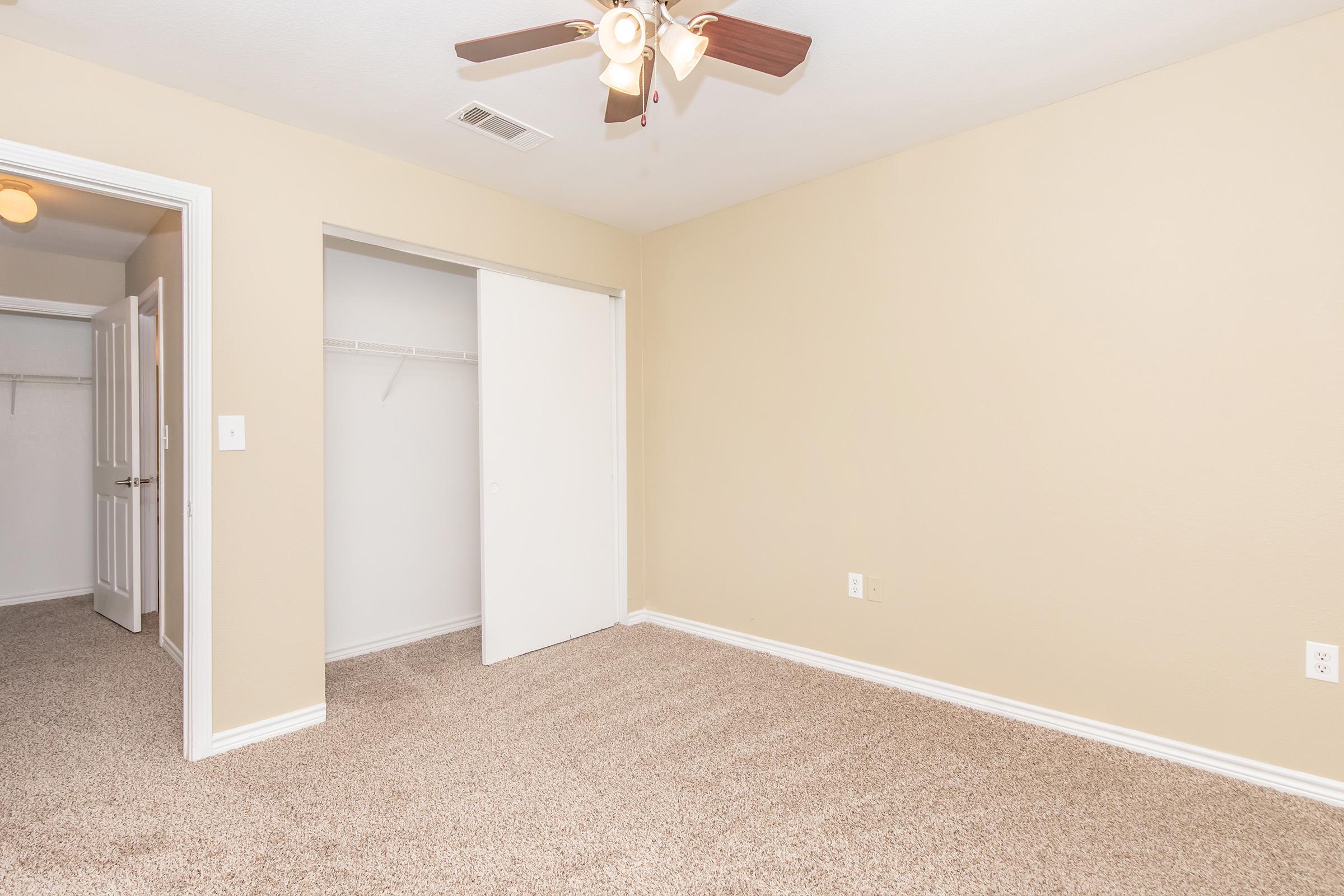 Empty room with beige walls and carpet, featuring a ceiling fan with wooden blades. A closet with sliding doors is visible on one side, and there is another door leading to a hallway. The space is well-lit with natural light.