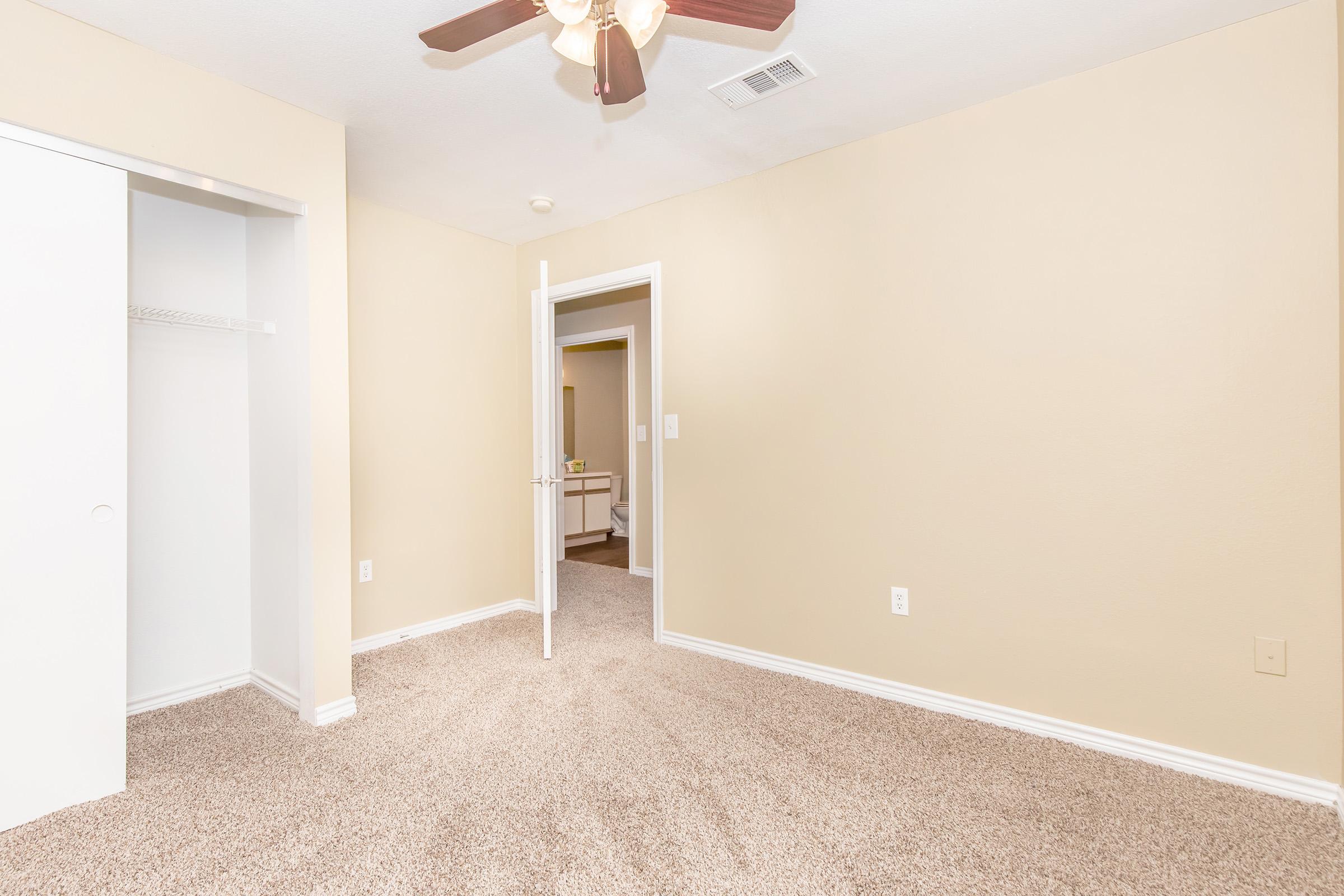A softly lit room with beige walls and carpet. It features a ceiling fan and an open doorway leading to a bathroom. A closet with a sliding door is visible on the left side, while the right side shows an additional open door. The space appears clean and well-maintained, conveying a warm and inviting atmosphere.