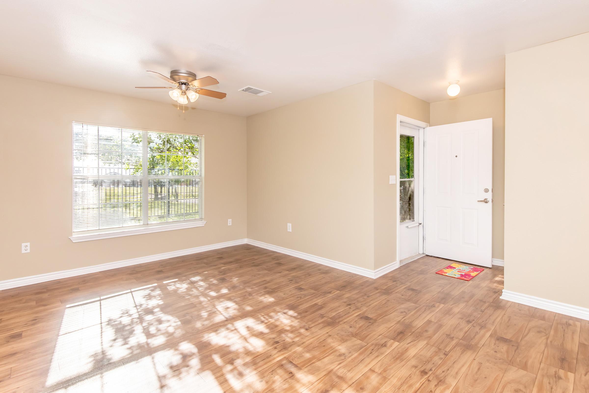A bright, empty room with light-colored walls and a wooden floor. A ceiling fan is mounted in the center, and natural light filters through a window with a view of greenery outside. A door with a small welcome mat leads to the entrance, creating a welcoming atmosphere.