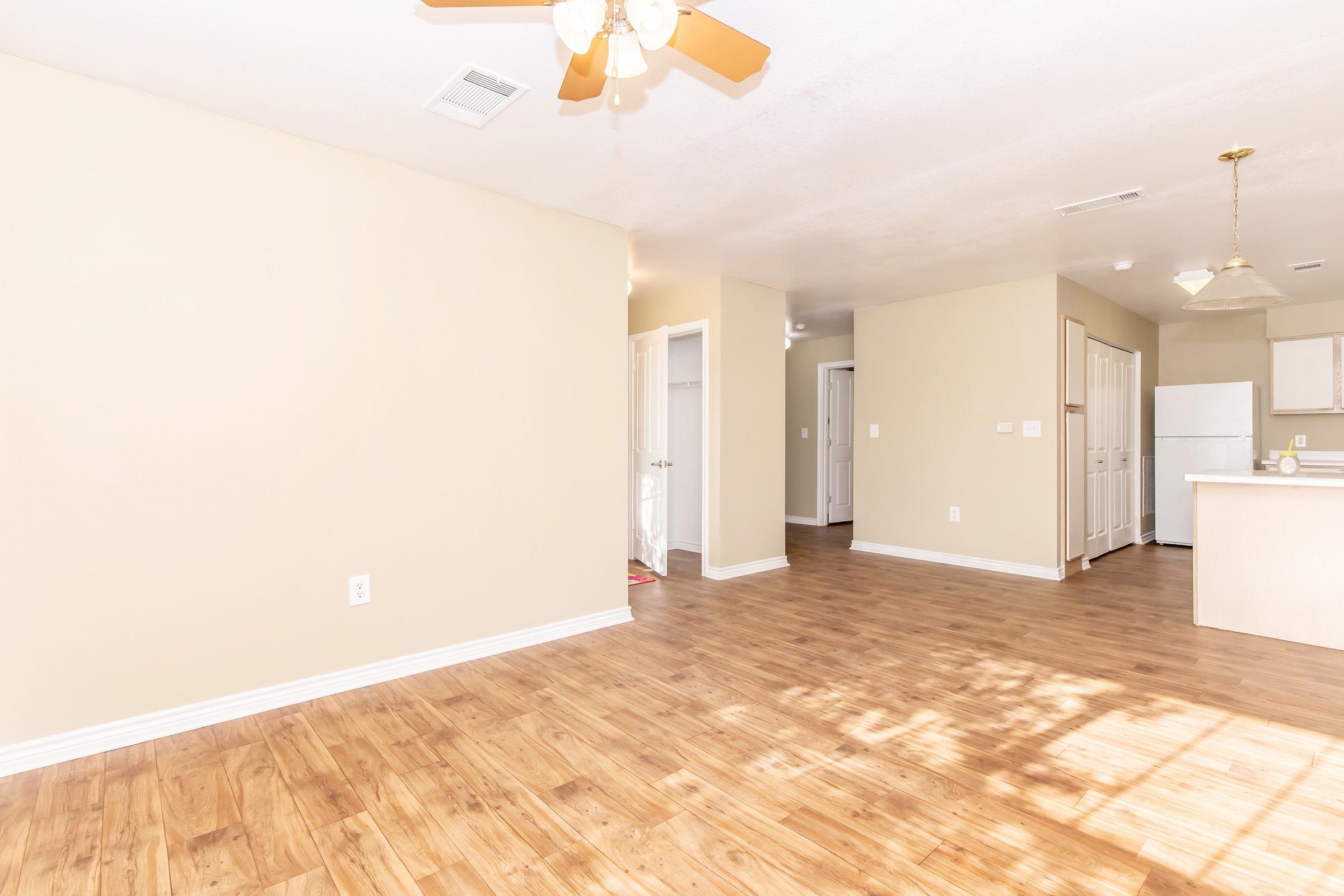 Spacious indoor area featuring wooden flooring, beige walls, and a ceiling fan. The room includes an open layout with a doorway leading to a kitchen that has white cabinets and appliances. Natural light brightens the space, creating an inviting atmosphere.