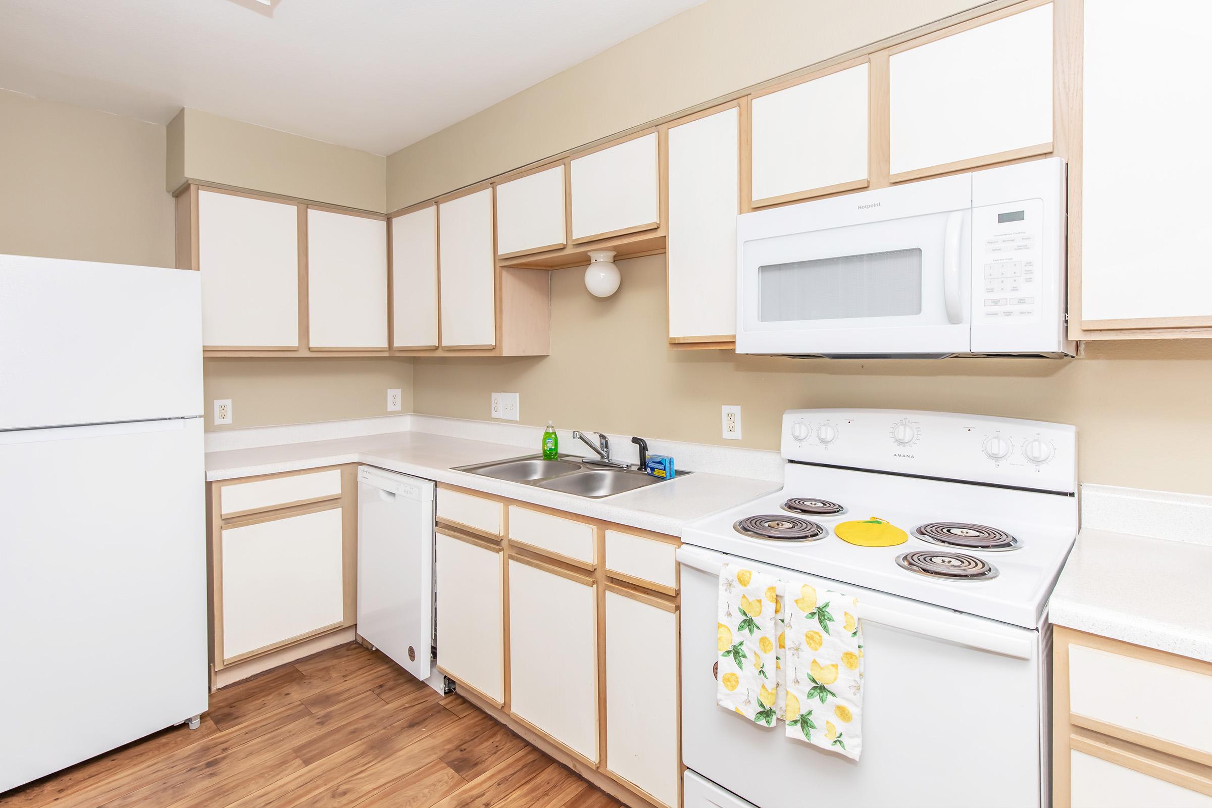 Bright, modern kitchen featuring white appliances, including a refrigerator and microwave, along with a white stove. Light-colored cabinets with a simple design line the walls, and a countertop is equipped with green dish soap and a yellow plate. A patterned dish towel hangs from the oven handle.