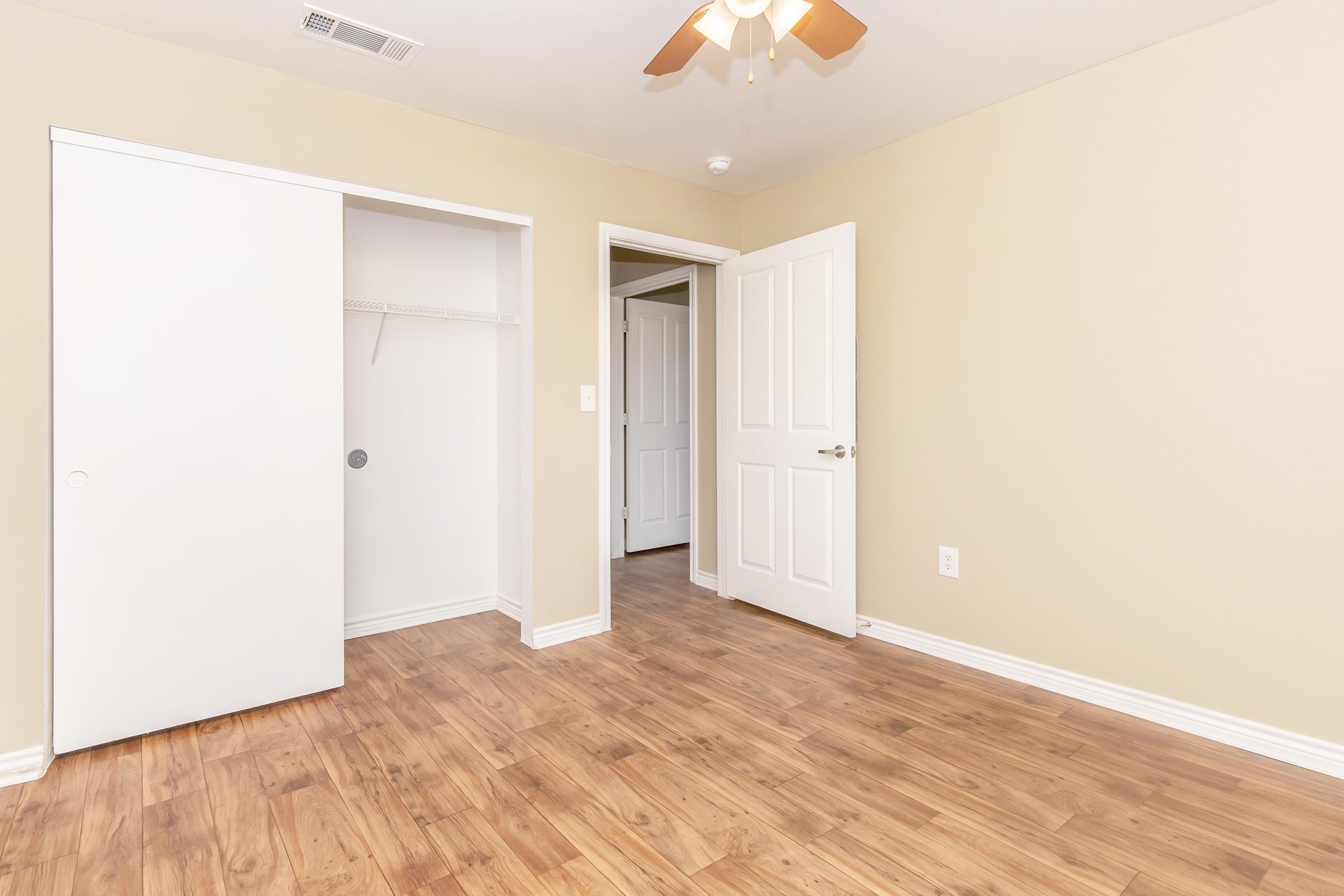 A neutral-colored room featuring light brown laminate flooring, a ceiling fan, and white doors. One door leads to a closet with sliding doors, while another door opens to a hallway. The walls are painted in a light shade, creating a bright and airy atmosphere.