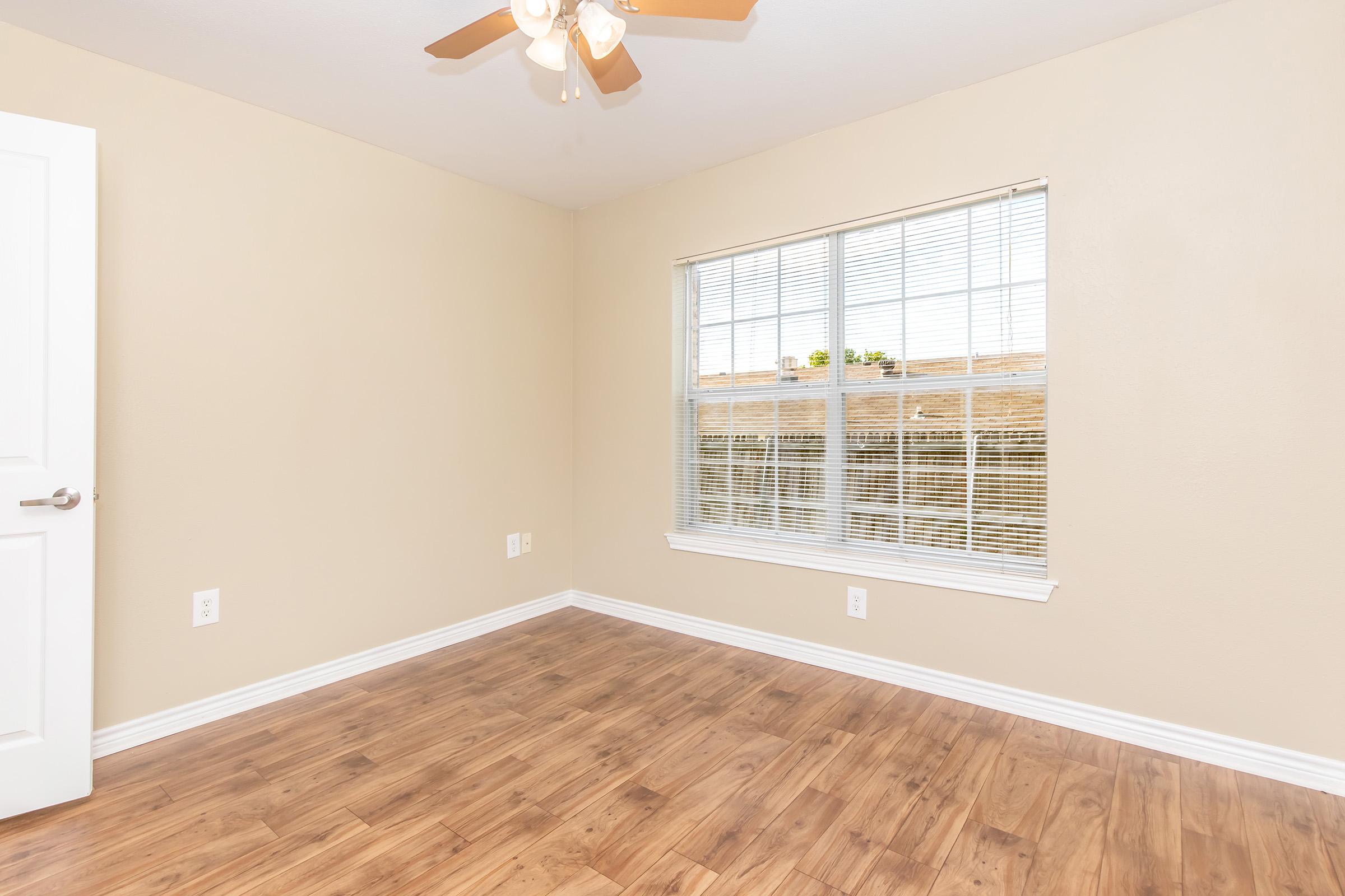 A well-lit empty room with beige walls and a ceiling fan. It features a large window with horizontal blinds, allowing natural light to enter. The flooring is light-colored wood, and there are no furnishings in the space. A closed white door is visible on the left.