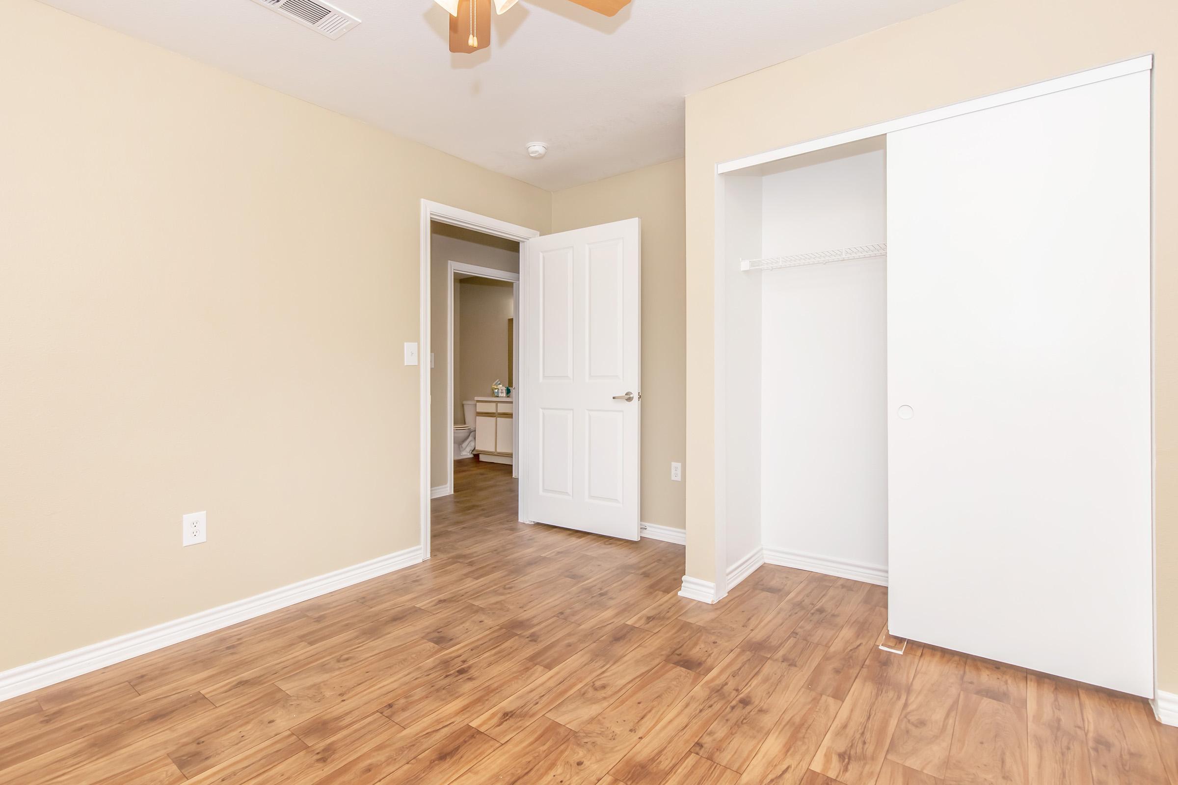 Interior view of a room with light beige walls and hardwood flooring. A closet with white doors is visible on the right side, and a doorway leads to another room in the background. Natural light is present, highlighting the clean and simple design.