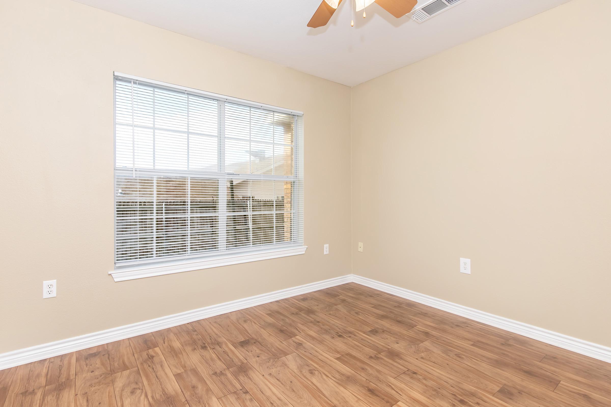 Interior view of a vacant room featuring a light beige wall and wooden flooring. A large window with white blinds allows natural light to enter the space, and a ceiling fan is mounted above. The room has no furniture, creating a clean and open appearance.