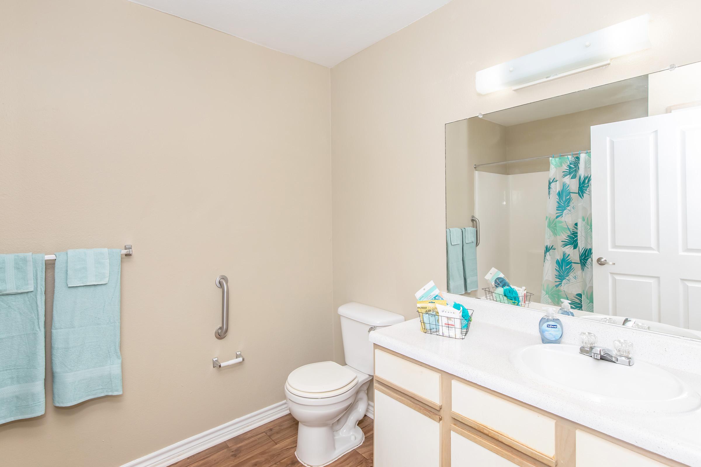 A clean, light-colored bathroom featuring a white toilet, a double sink with a light countertop, and a large mirror above it. The walls are painted beige, and there's a shower curtain with a tropical pattern. Two aqua towels hang on a rack, and a small basket with toiletries is on the counter.