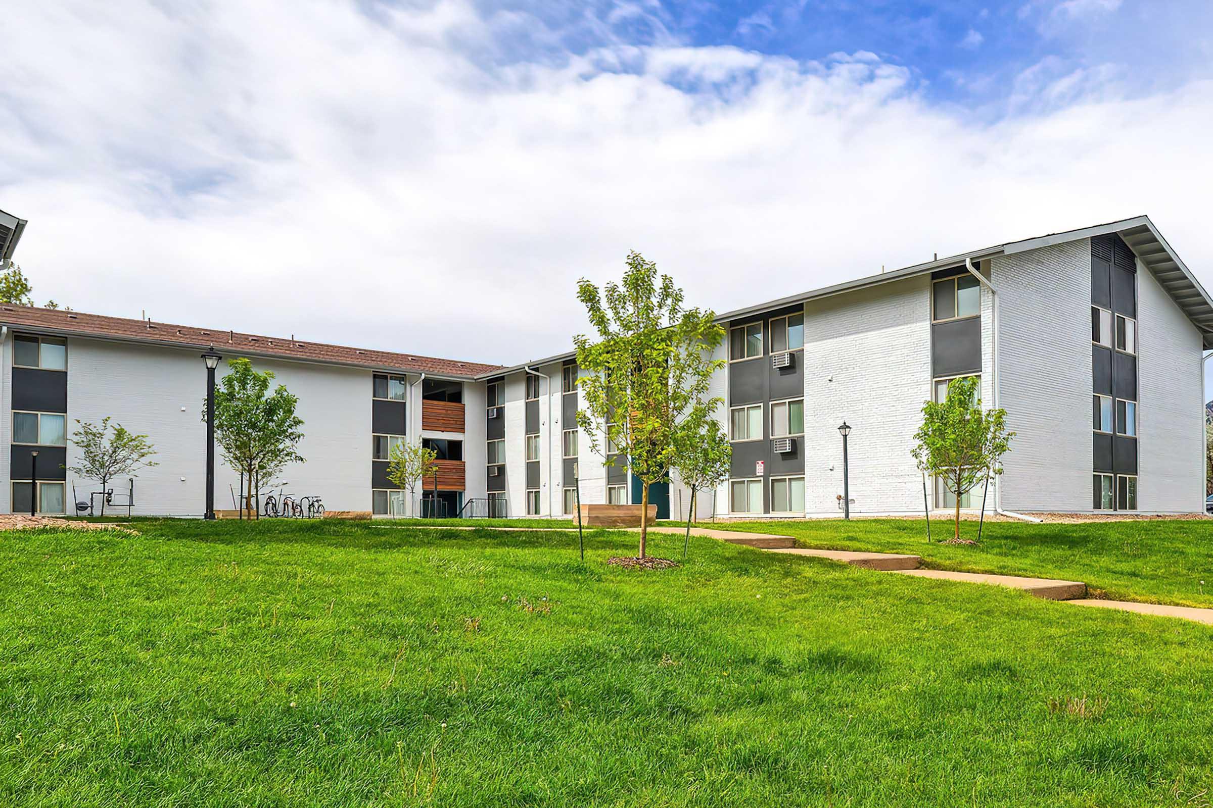 A modern apartment building with a landscaped green area in front. There are young trees and a pathway leading to the entrance. The building has a mix of brick and white siding, with windows that reflect the sky. The scene is set under a partly cloudy sky.