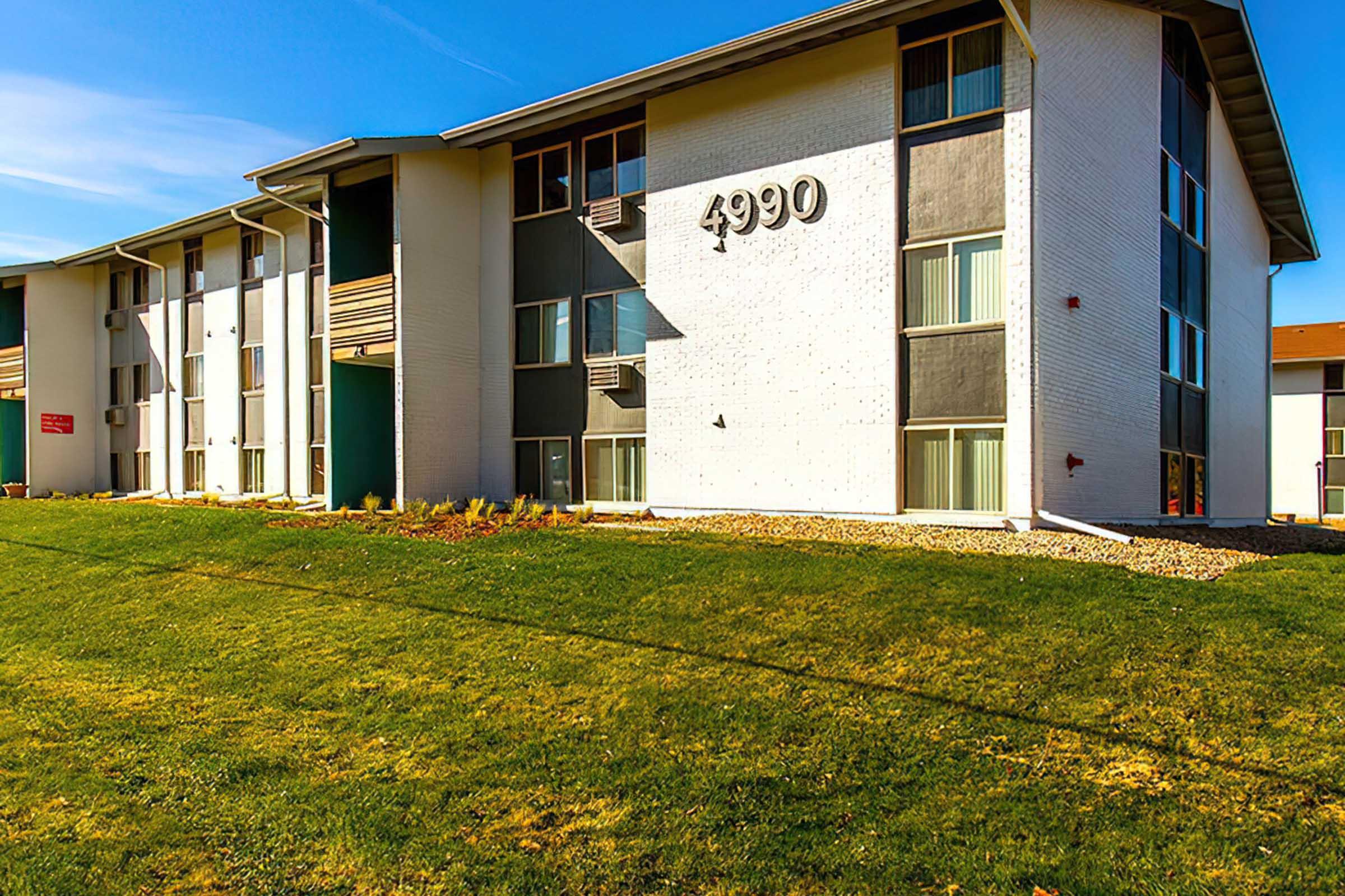 A modern multi-story apartment building featuring a green lawn in the foreground. The building has large windows, a mix of light and dark exterior colors, and the number "4990" prominently displayed on the front facade. The sky is clear and blue, suggesting a sunny day.