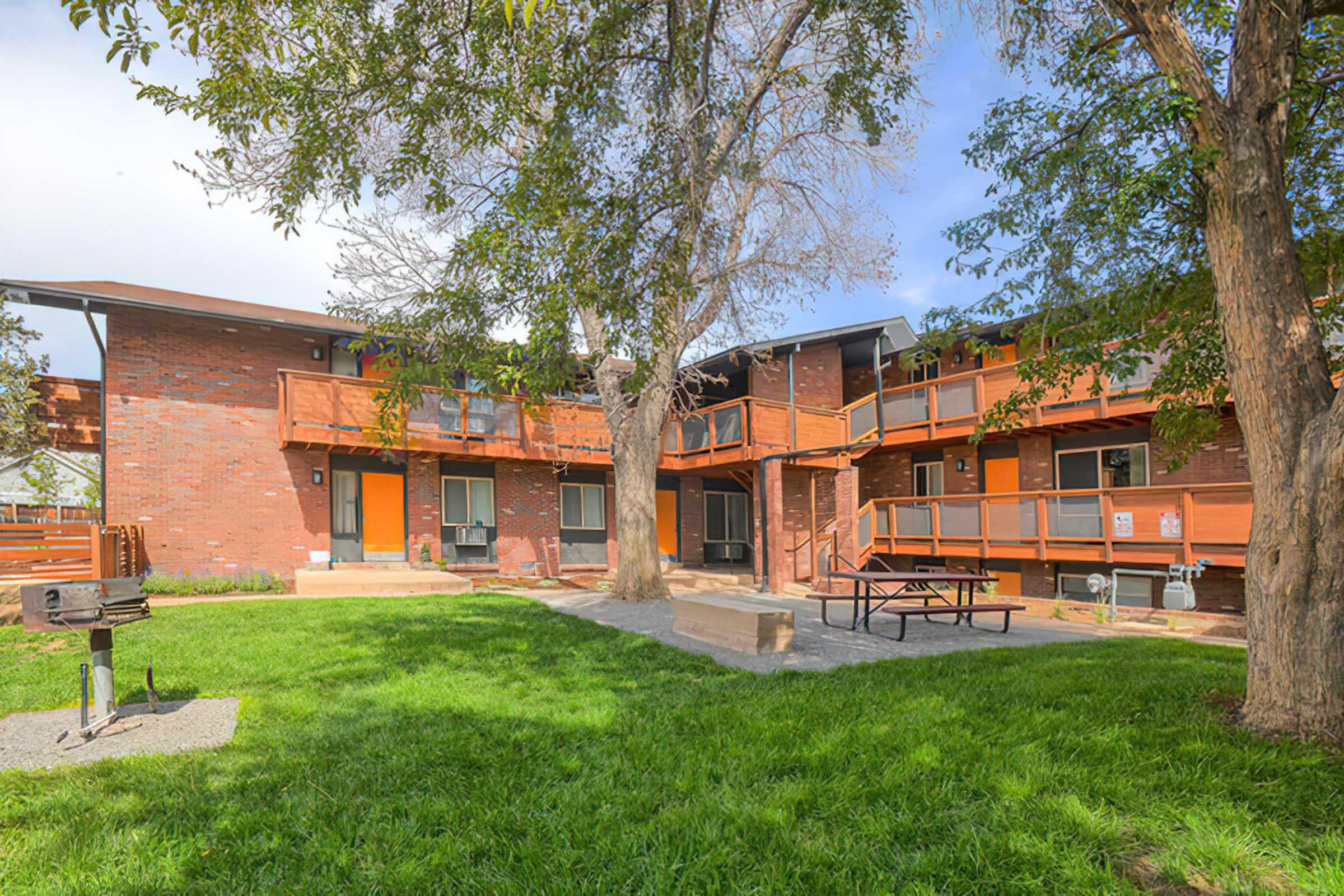 A grassy courtyard features several apartment buildings with red brick exteriors and wooden balconies. There is a picnic table and grill area on the lawn, surrounded by trees, creating a welcoming outdoor space for residents.
