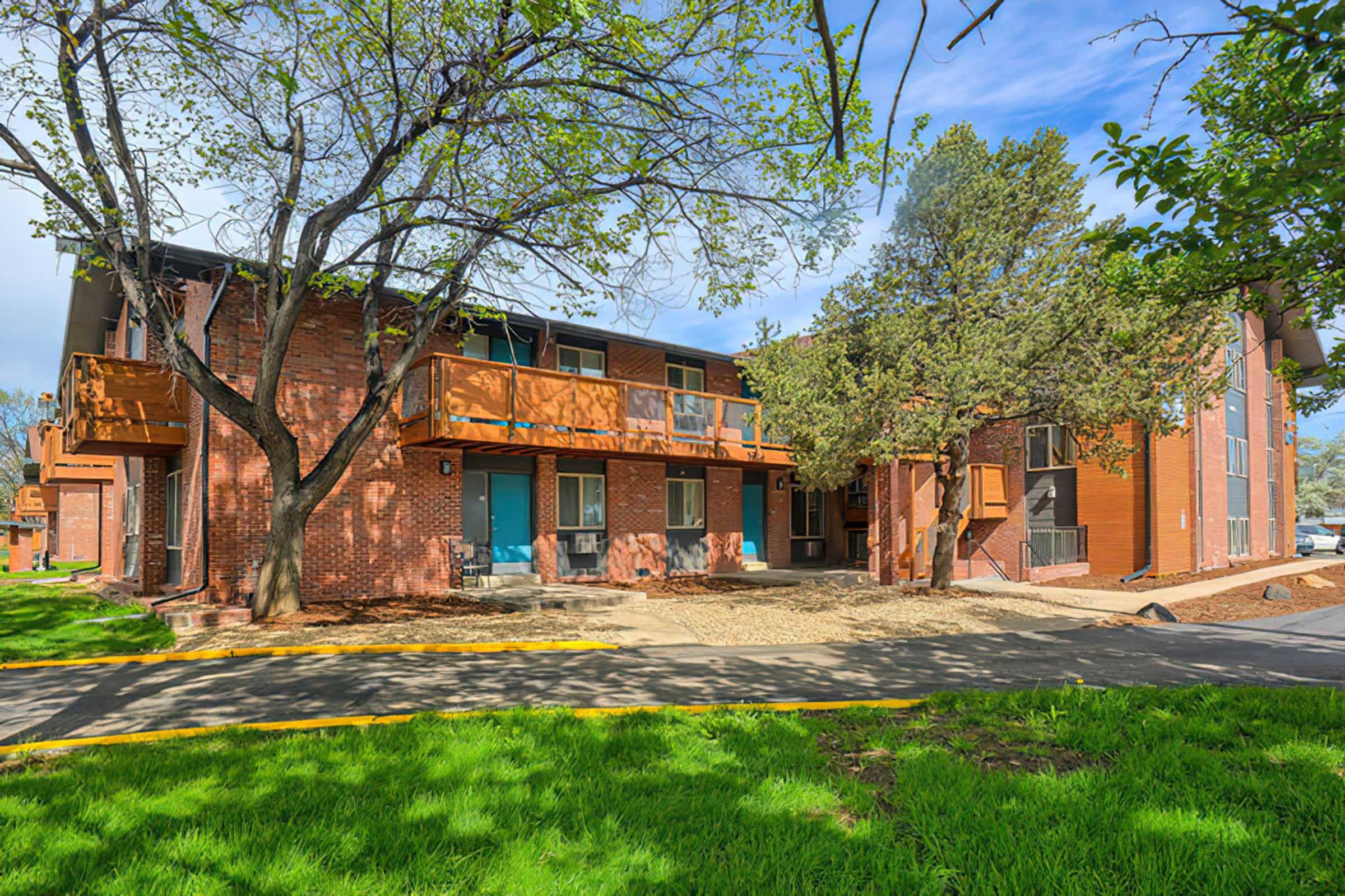 A view of a two-story brick apartment building surrounded by green grass and trees. The building features outdoor balconies and is set against a clear blue sky. A paved walkway leads up to the entrance, adding to the inviting atmosphere of the residential area.