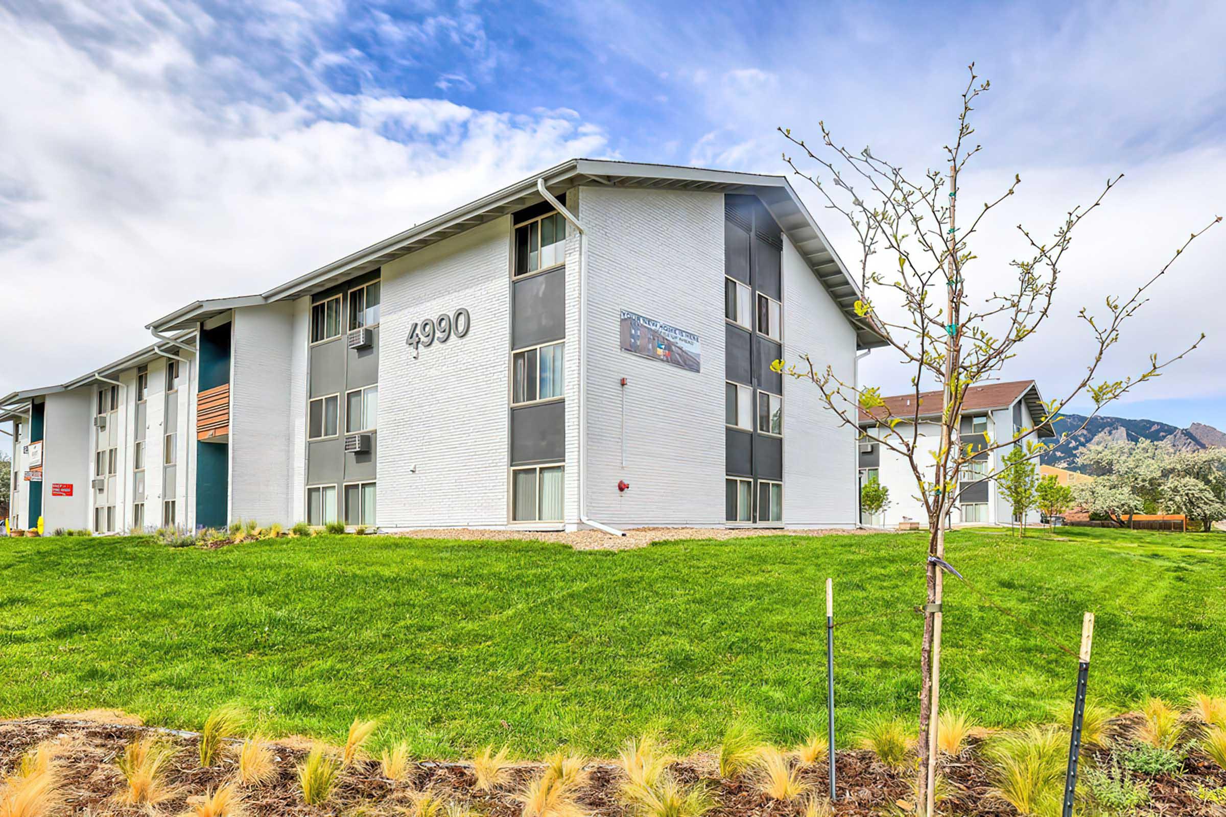Three-story white apartment building with the number "4990" prominently displayed on the side. The building is surrounded by green grass and small trees, with mountains visible in the background under a partly cloudy sky.