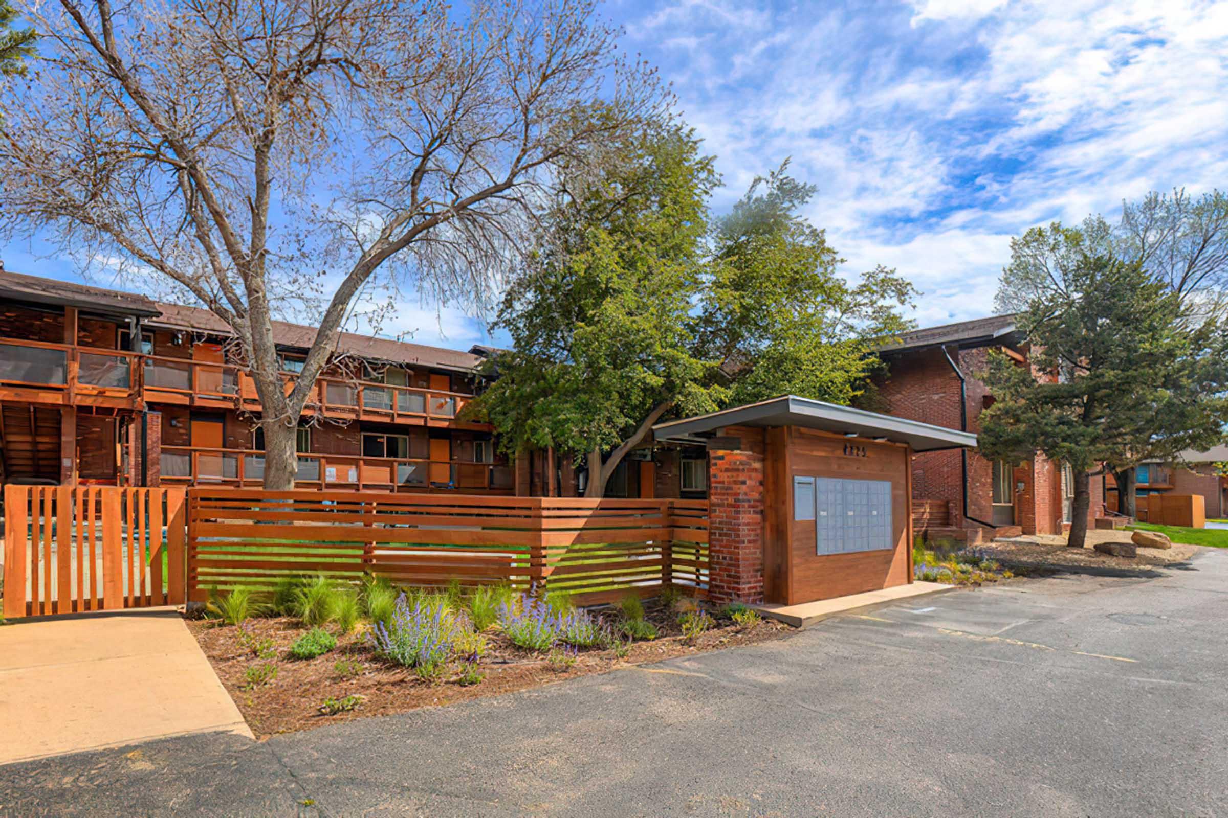 A modern multi-unit apartment building featuring wooden accents and greenery. There are trees and shrubs in the landscape, with a wooden fence in the foreground. The sky is partly cloudy, adding to the appealing outdoor setting.