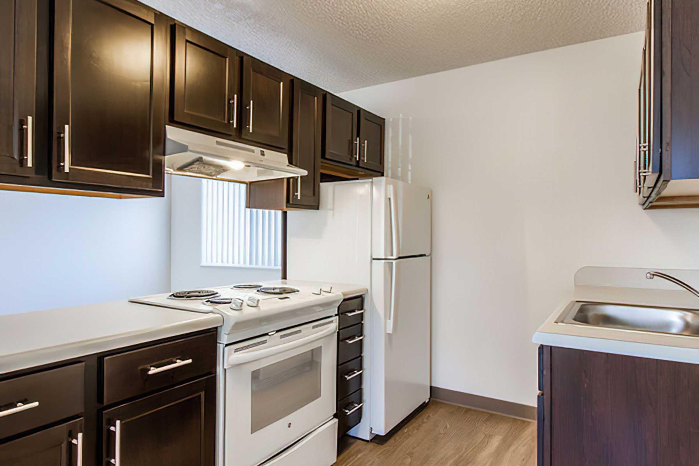 Modern kitchen featuring dark wood cabinets, a white stove and refrigerator, and a stainless steel sink. The layout includes a small countertop area and a window allowing natural light. The flooring is a light wood laminate, creating a warm and inviting atmosphere.
