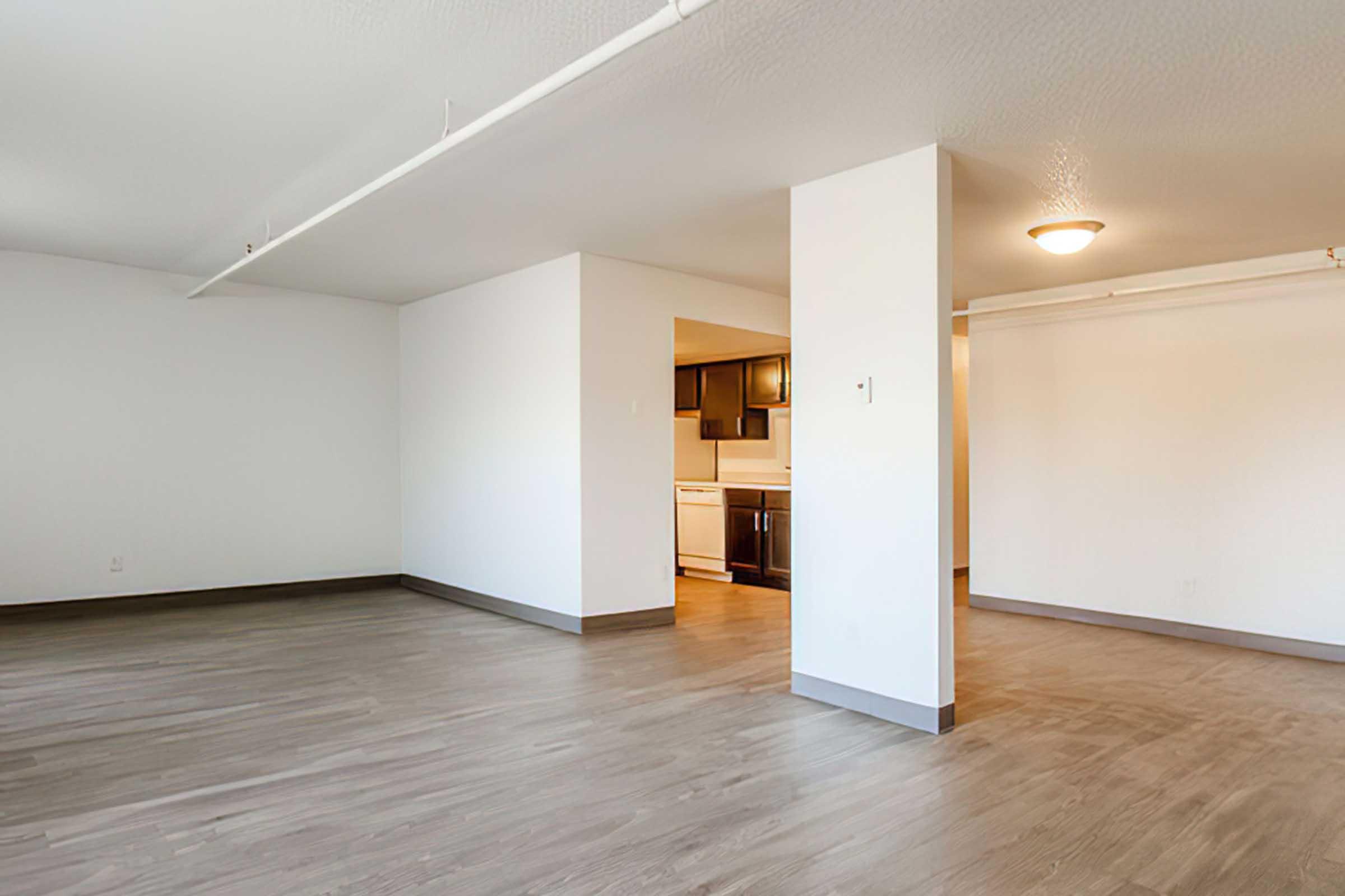 An empty room with light-colored walls and a hardwood floor. There is a doorway leading to a kitchen area in the background with dark cabinets. The ceiling has a light fixture, and there are no furnishings present in the space, creating a clean, minimalist look.