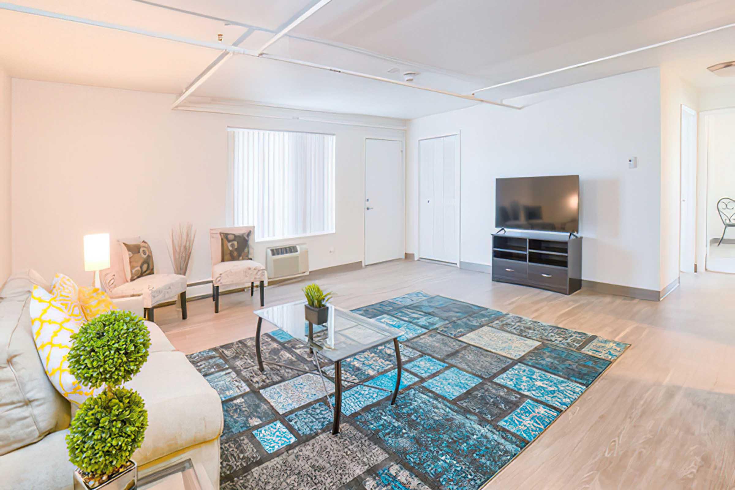 Bright and spacious living room featuring a light-colored sofa with decorative pillows, a coffee table on a patterned rug, and a flat-screen TV on a stand. Natural light pours in through a window, enhancing the modern decor with plants and minimalist furniture.