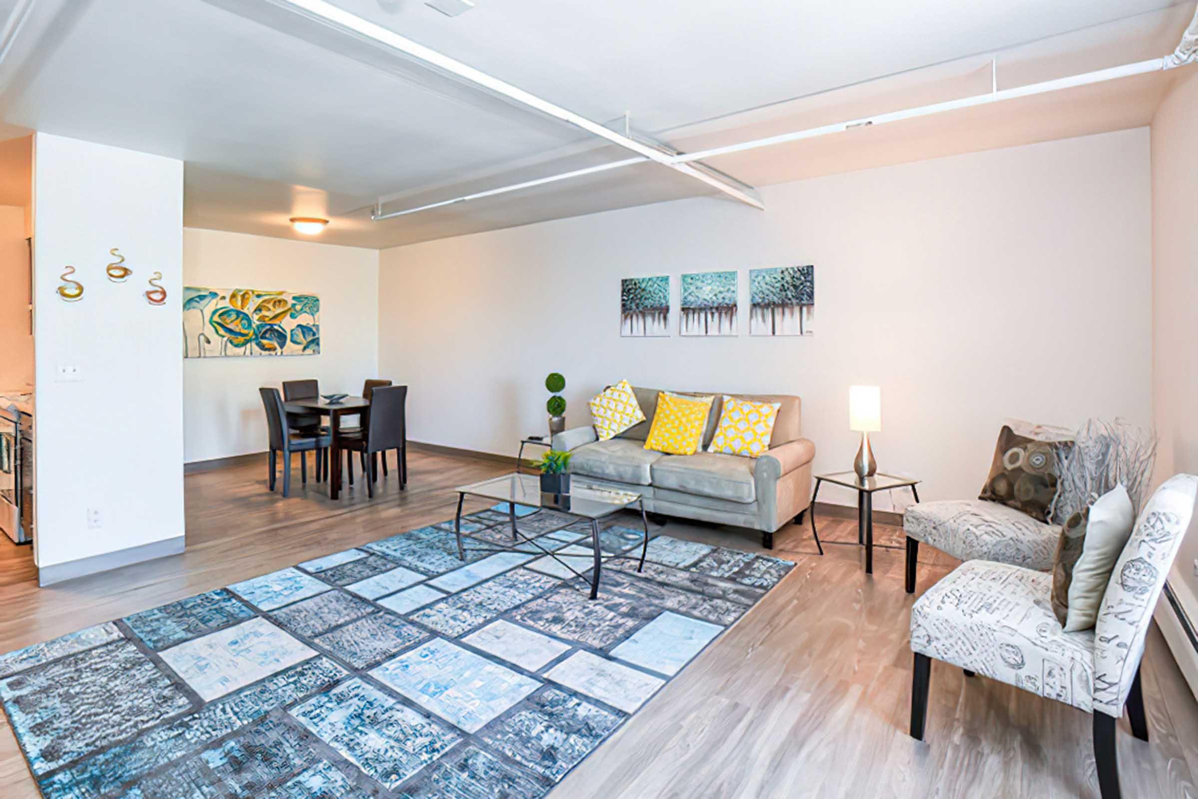 A modern, well-lit living room featuring a gray couch with colorful throw pillows, a glass coffee table, and a patterned area rug. There is a small dining area with a table and chairs in the background, along with vibrant wall art depicting abstract landscapes.