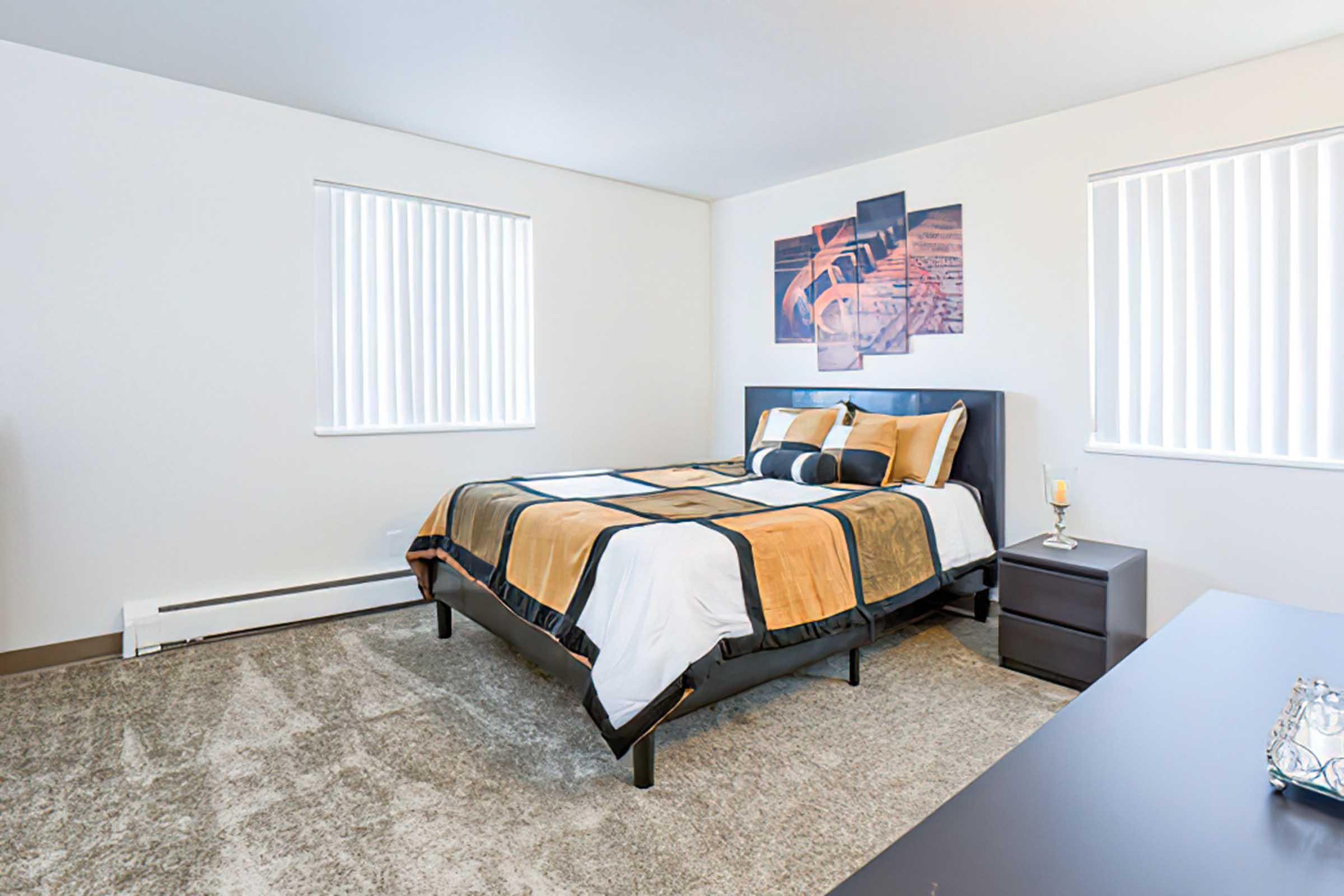 A modern bedroom featuring a neatly made bed with a black and gold comforter, two window treatments allowing natural light, and a nightstand with a candle. The walls are painted white, and there's a minimalist aesthetic with neutral-colored carpeting and a stylish wall art piece above the bed.