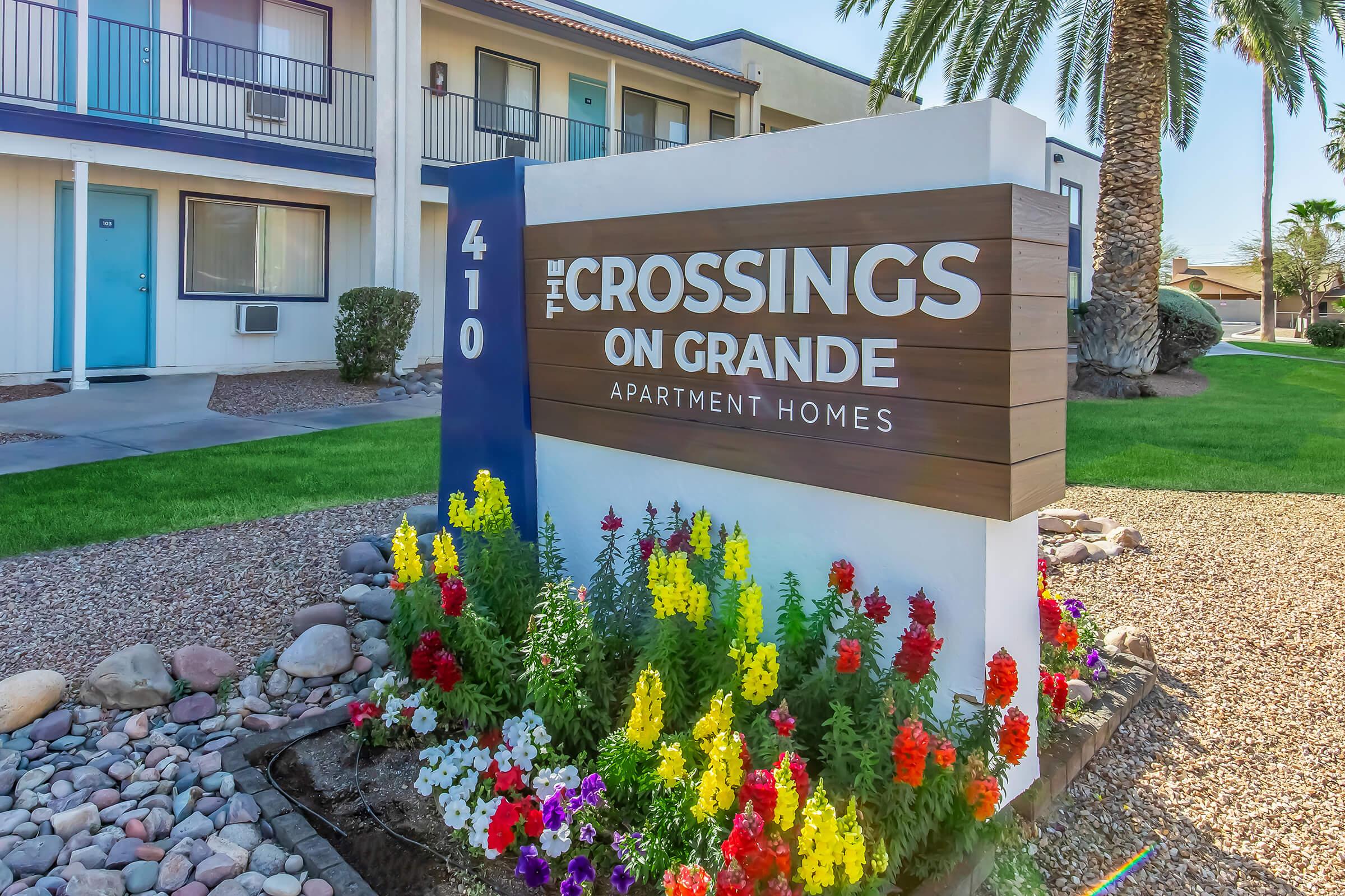Sign for The Crossings on Grande Apartment Homes, featuring the address 410. The sign is surrounded by colorful flowers and landscaped greenery. In the background, there are buildings with balconies and palm trees, indicating a well-maintained apartment complex.