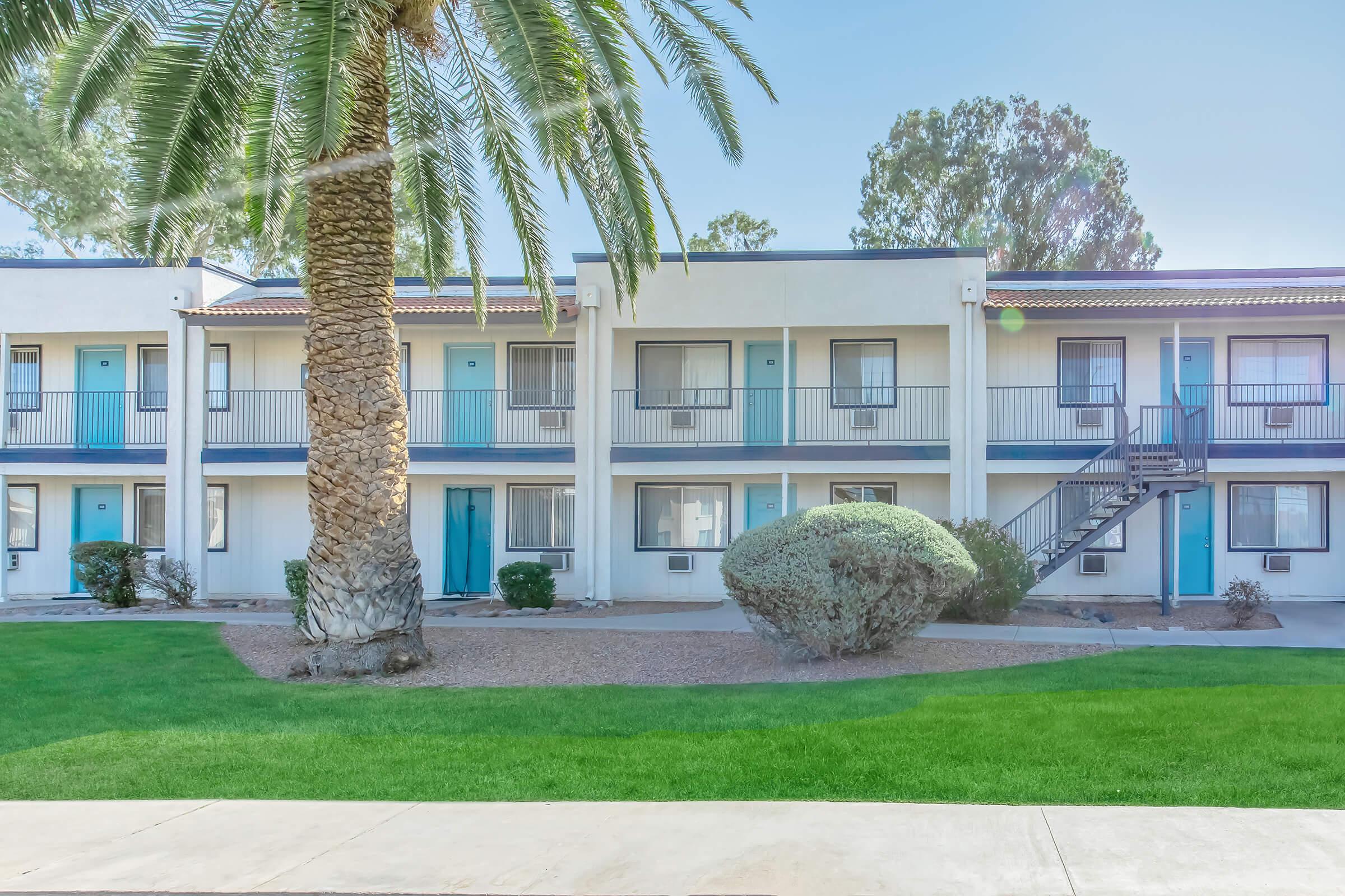 A two-story building with multiple rooms, featuring blue doors and balconies, surrounded by green grass and a neatly trimmed bush. A tall palm tree is in the foreground, and a metal staircase leads to the upper floor. The setting appears sunny and well-maintained.