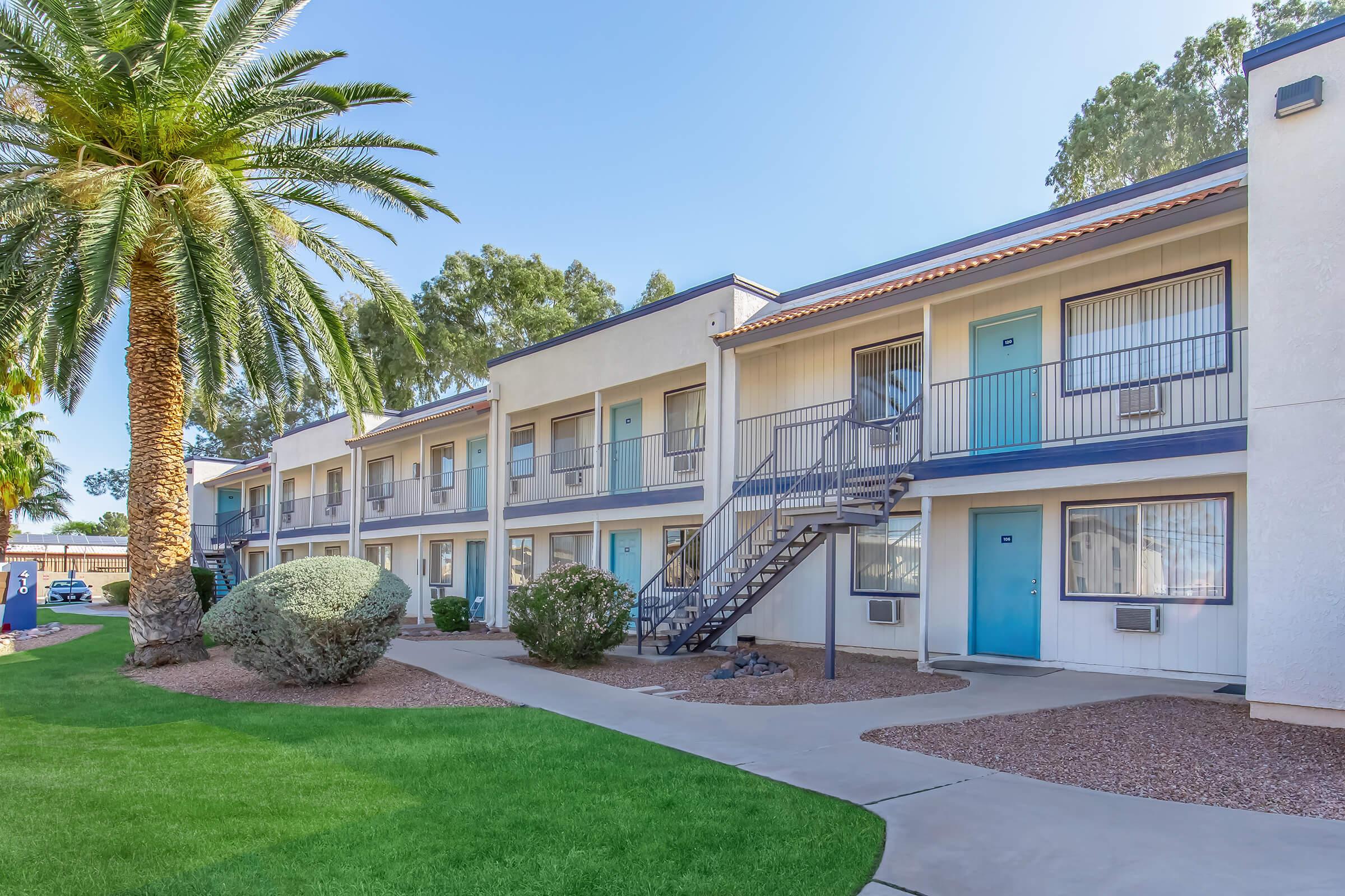 A view of a two-story apartment building featuring blue doors and a staircase, surrounded by manicured grass and palm trees. The building includes several windows, air conditioning units, and a neatly landscaped area with bushes.