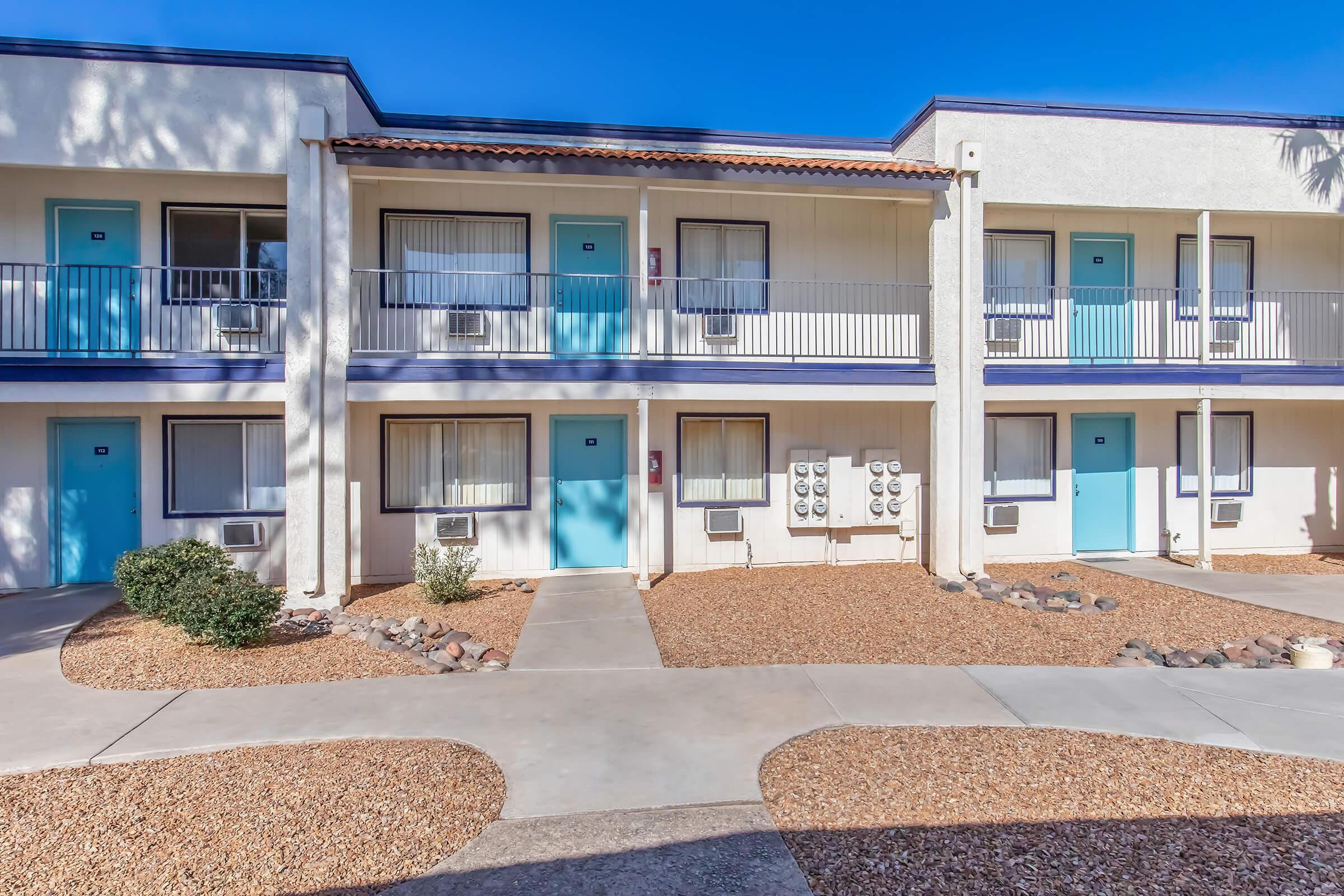 A view of a two-story motel with several rooms featuring blue doors and balconies. The courtyard in front is landscaped with gravel, small shrubs, and rocks, surrounded by sunny skies. Air conditioning units are visible beneath each window, giving the impression of a warm climate.