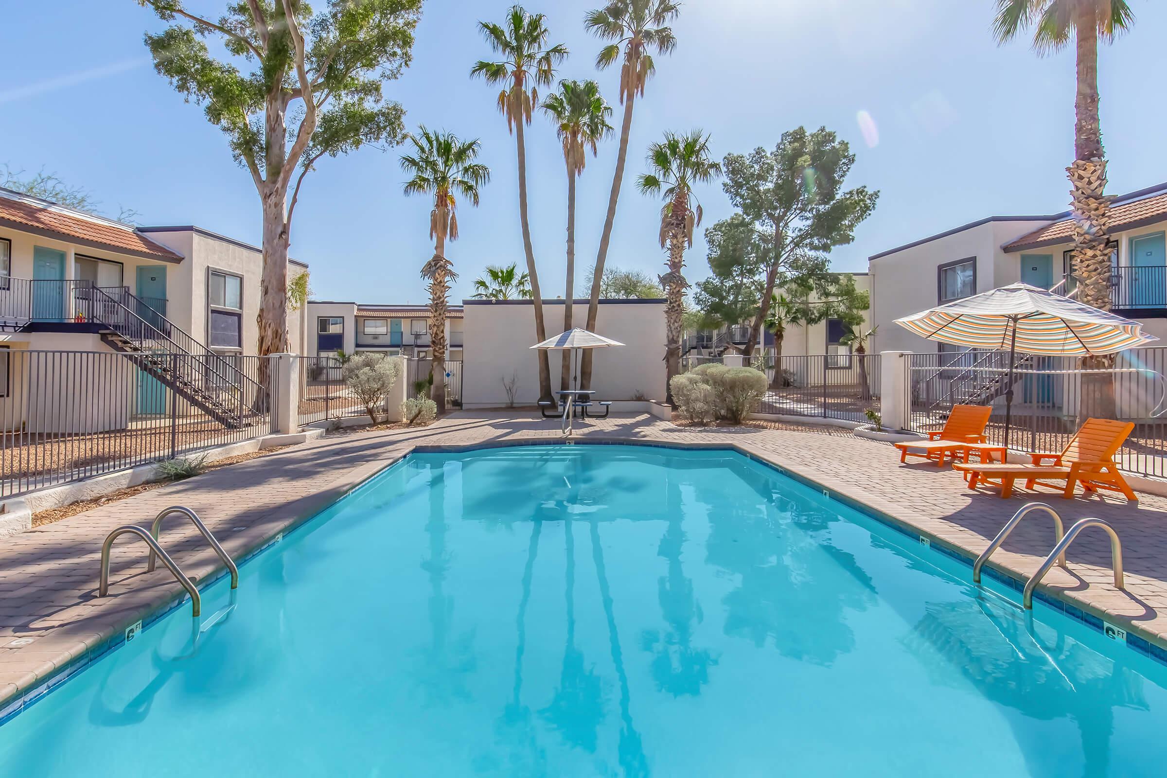 A bright outdoor swimming pool surrounded by palm trees and lounge chairs. Umbrellas provide shade near the pool area, with apartment buildings in the background. The scene is under a clear blue sky, suggesting a warm and inviting atmosphere.