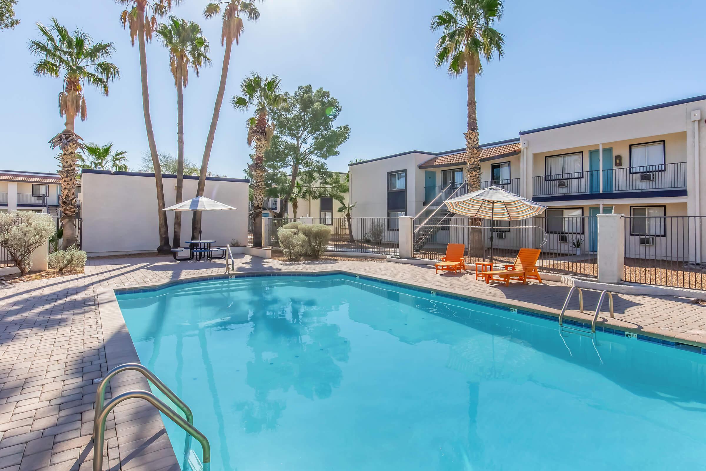 A clear, blue swimming pool surrounded by palm trees and a sunny sky. Lounge chairs and an umbrella are positioned near the pool, with a small table nearby. Multi-story buildings with balconies are visible in the background, creating a relaxed outdoor atmosphere.