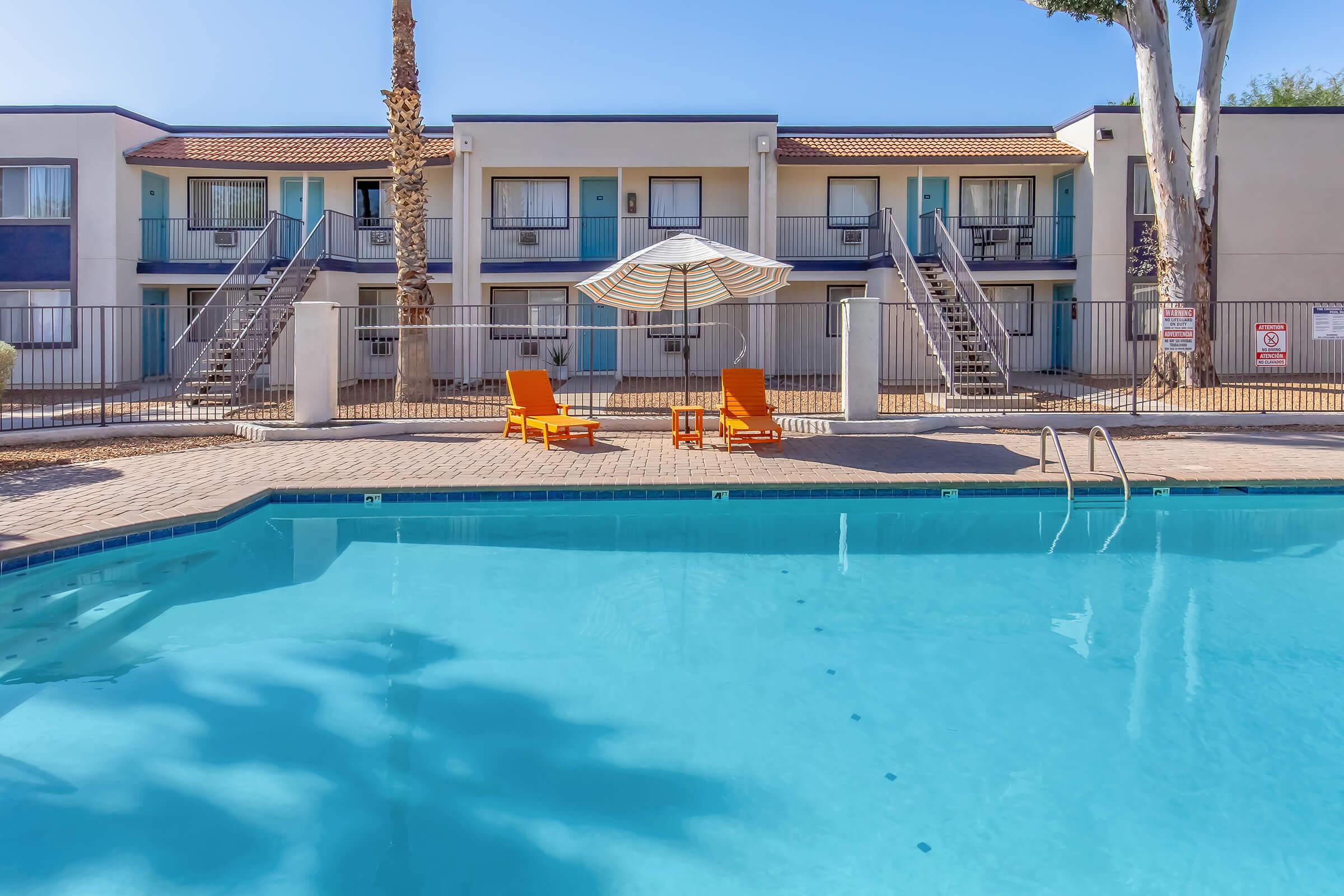 A swimming pool with clear blue water and orange lounge chairs beneath a striped umbrella. In the background, there are two-story apartment buildings with balconies and palm trees. The area is well-lit and inviting, creating a relaxing atmosphere.