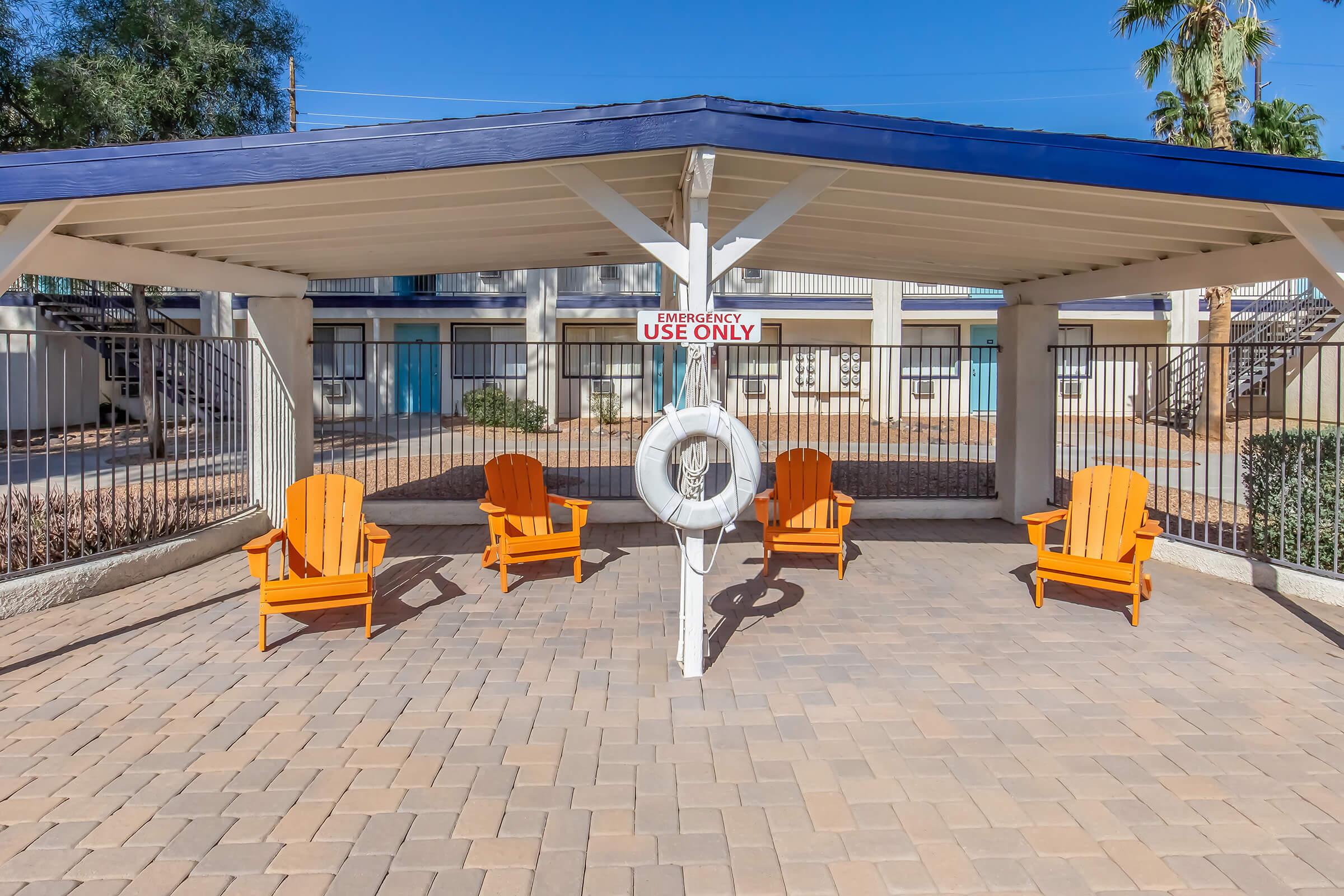 A covered area with four orange Adirondack chairs arranged around a central post holding a life ring. The backdrop features a fenced pool area with palm trees and buildings in the distance under a clear blue sky. A sign above the chairs reads "EMERGENCY USE ONLY."