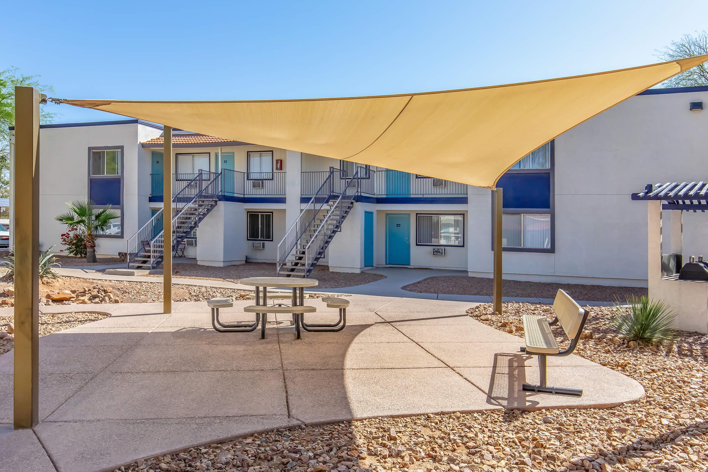A shaded outdoor seating area with a light-colored canopy over a round picnic table and chairs, surrounded by landscaped gravel and small plants, near an apartment building with blue accents and staircases. The scene is set in a sunny environment.