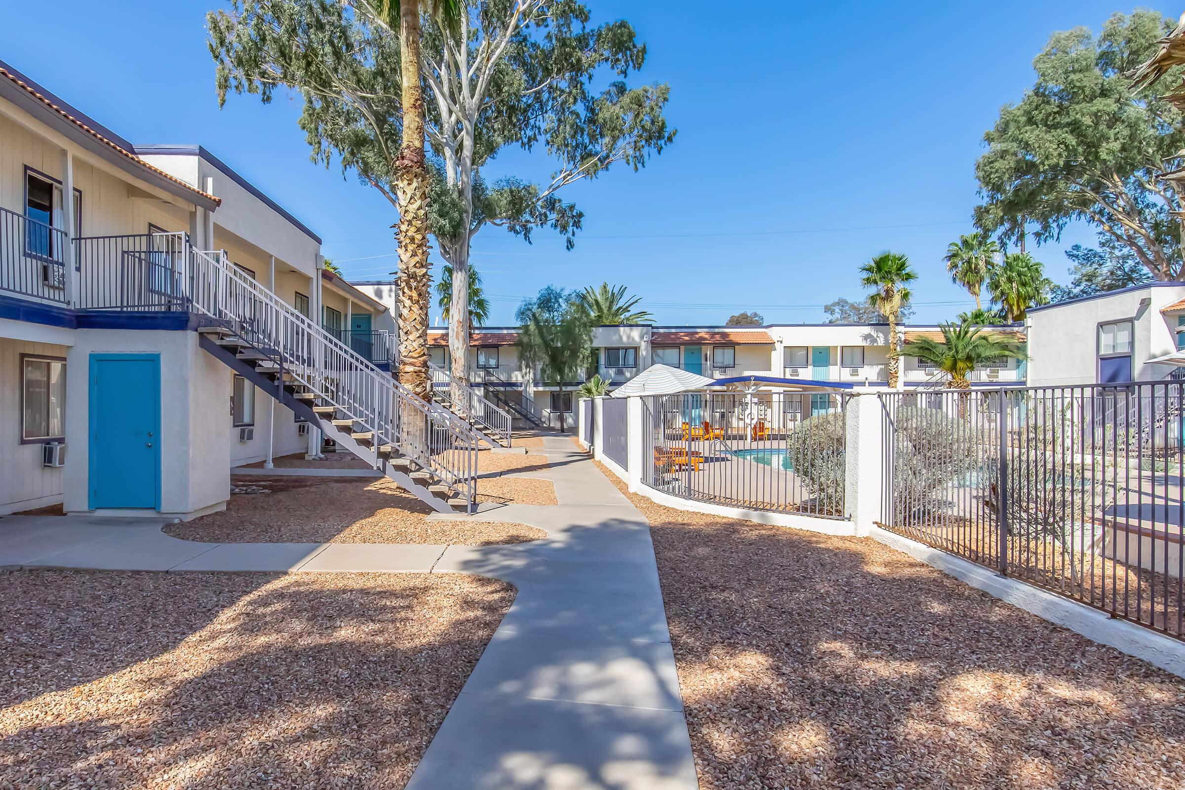 A courtyard view of a multi-unit residential building with a walkway, palm trees, and a swimming pool in the background. The area is landscaped with gravel and features a staircase leading to upper levels. Bright blue sky enhances the inviting atmosphere.