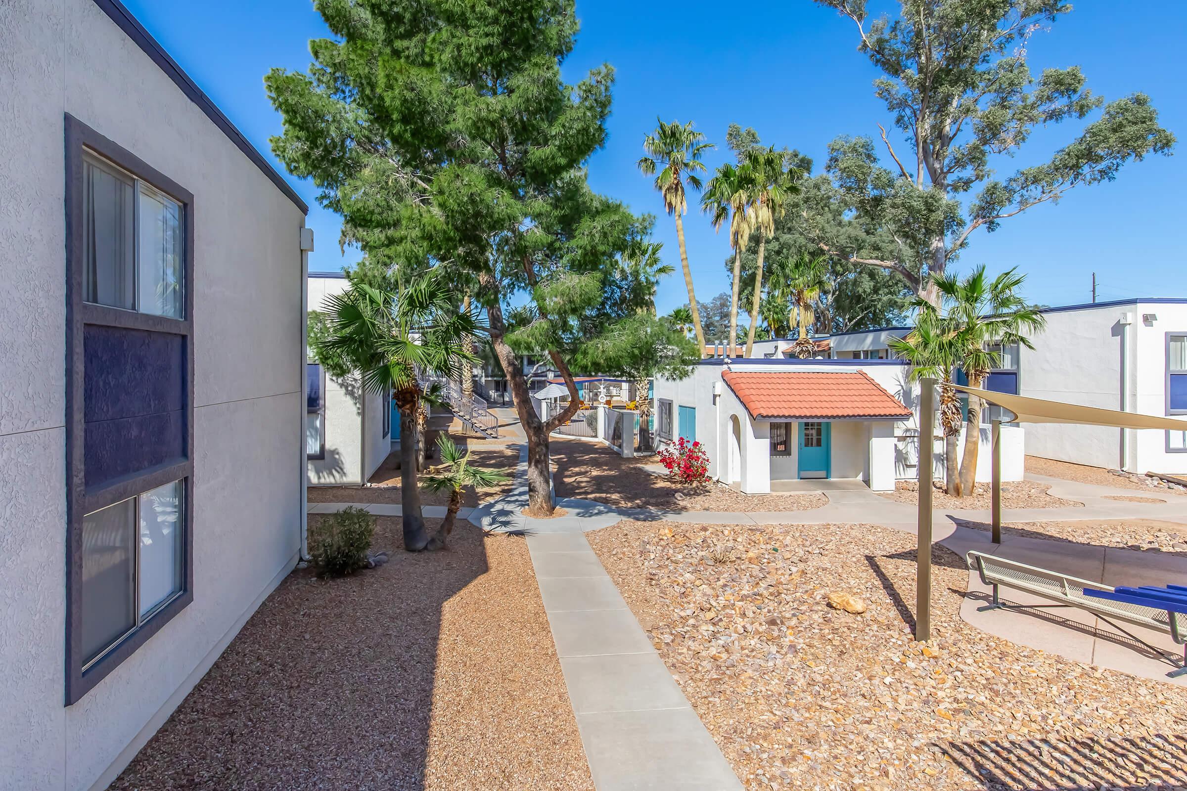 A sunny outdoor view of a residential complex featuring a pathway lined with palm trees and shrubs. The scene includes several white buildings, landscaped areas with rocks, and a glimpse of colorful flowers. It's a clear day with blue skies, creating a vibrant and welcoming atmosphere.