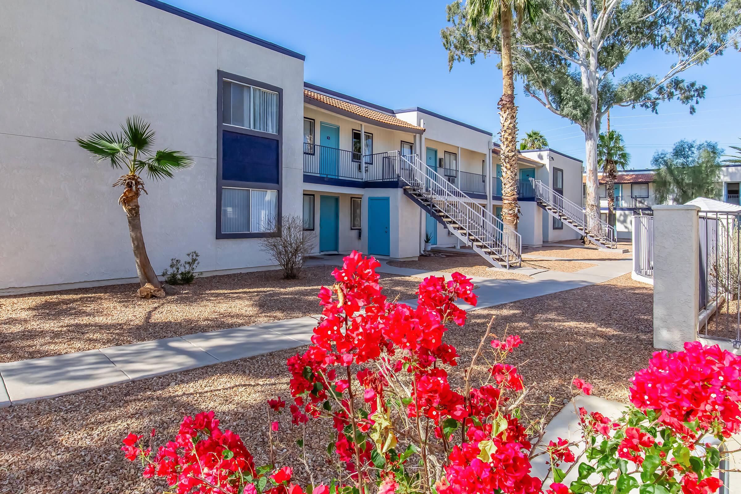 A view of a two-story apartment building surrounded by a landscaped area with blooming red flowers in the foreground. Palm trees and other greenery are visible alongside a gravel walkway leading to the entrance of the apartments. Clear blue skies provide a bright backdrop.